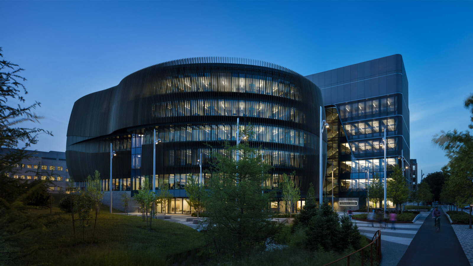 東北大學創新 ISEC 大樓 壁紙 — 一座現代弧形玻璃建築在暮色天空下溫暖發光，周圍有樹木和鋪砌的人行道。