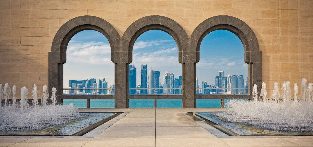 卡塔爾古 Islamic 建築 壁紙 — 三個石拱門框住現代城市天際線，下方是碧綠海水，前景有噴泉噴灑。
