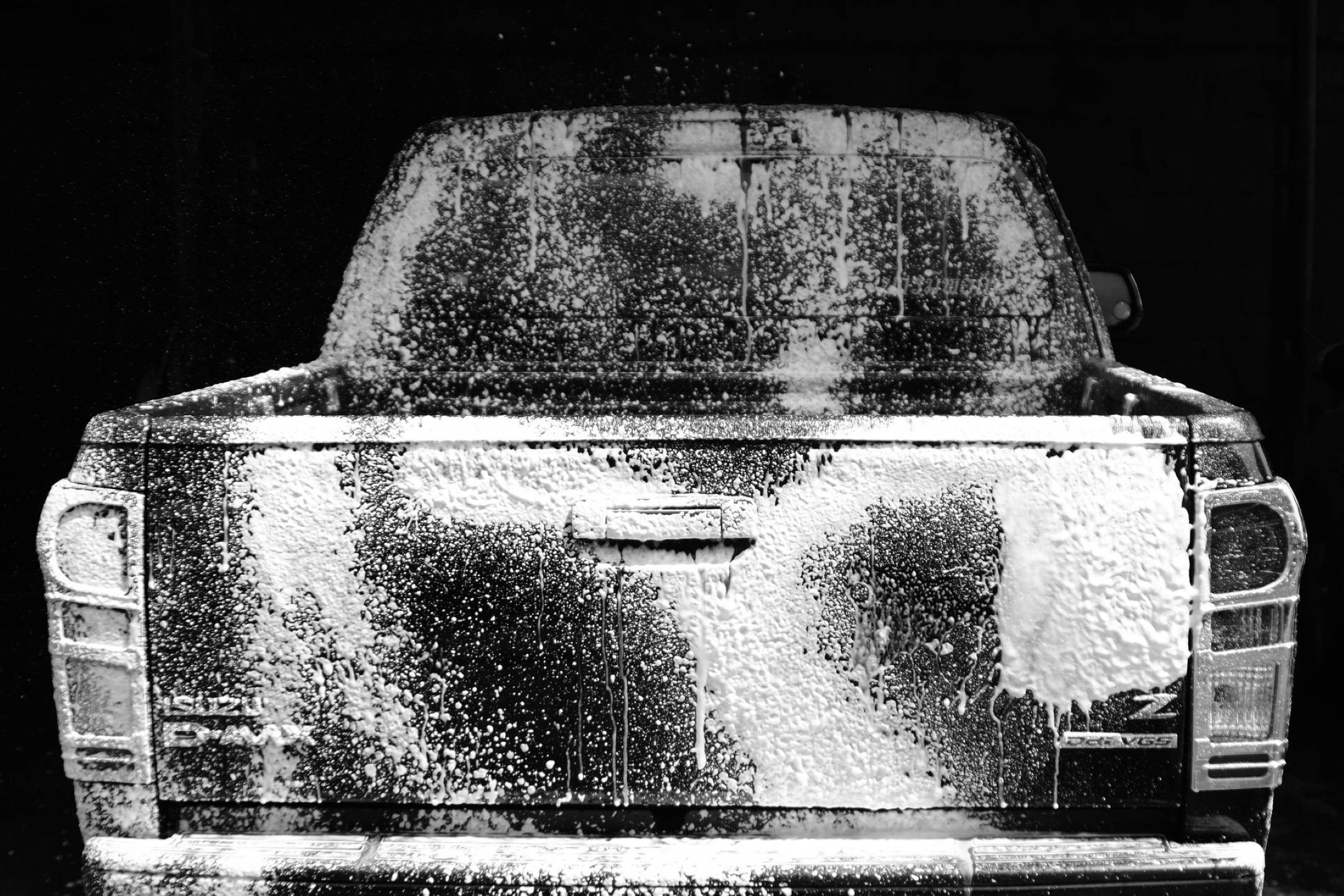 Isuzu D-Max皮卡車洗車場 壁紙 — 一張黑白照片，拍攝一輛皮卡車的後部，覆蓋著厚厚的肥皂泡沫，背景為深色。