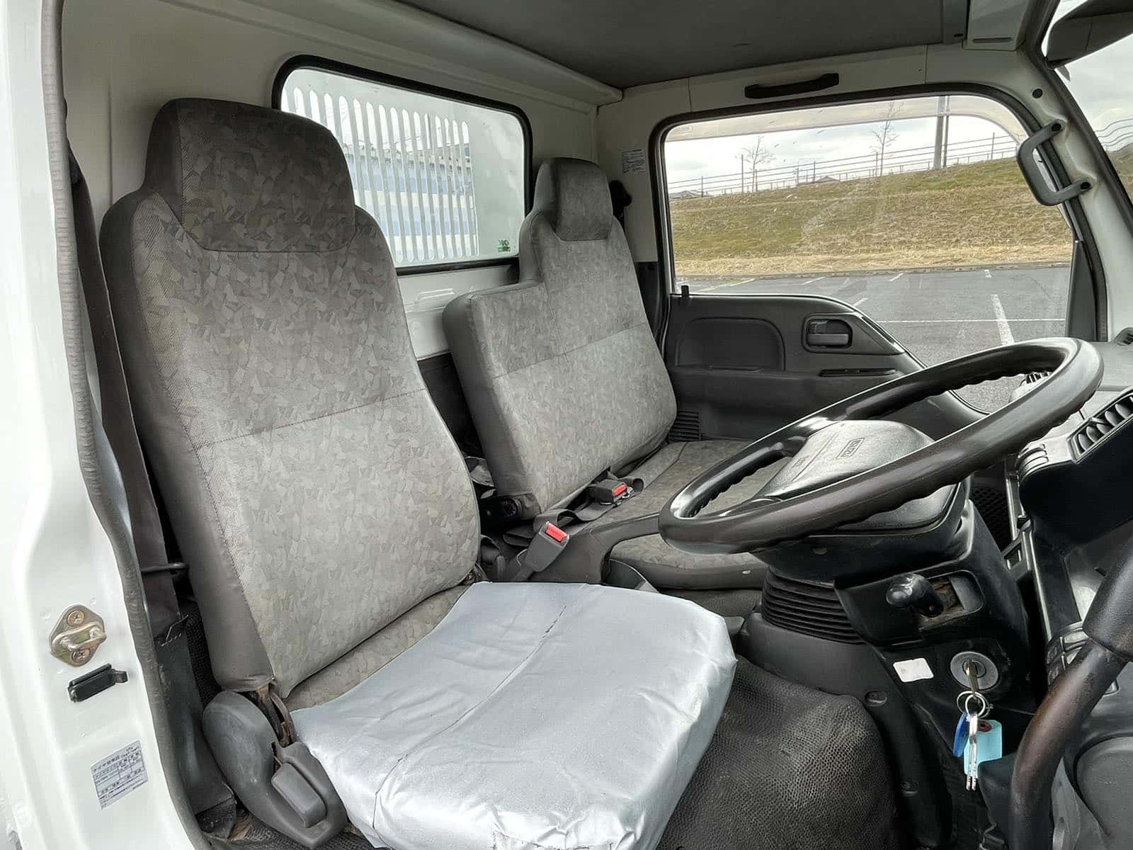 Isuzu Elf 卡車駕駛艙內部 壁紙 — 白色客貨車內部，顯示灰色圖案座椅，其中一個用塑膠覆蓋，方向盤和儀表板上有鑰匙插入點火開關。