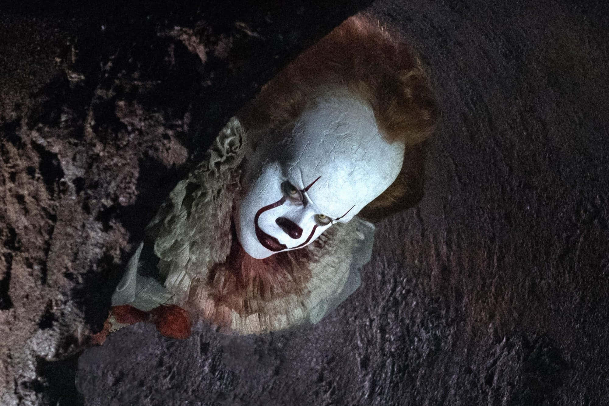 電影 IT 中 Pennywise 恐怖小丑圖片 壁紙 — 一個帶有白色臉龐、紅色嘴唇和橙色頭髮的威脅性小丑從黑暗的岩石裂縫中窺視。