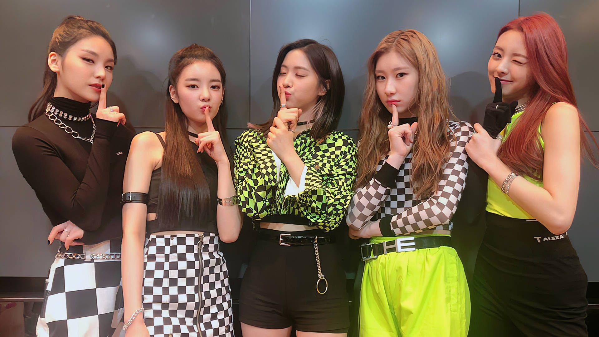 ITZY 成員出道時期 壁紙 — 五位時尚女性穿著大膽的格仔和螢光服裝，在黑暗的攝影棚牆前擺出手指放在嘴唇上的姿勢。