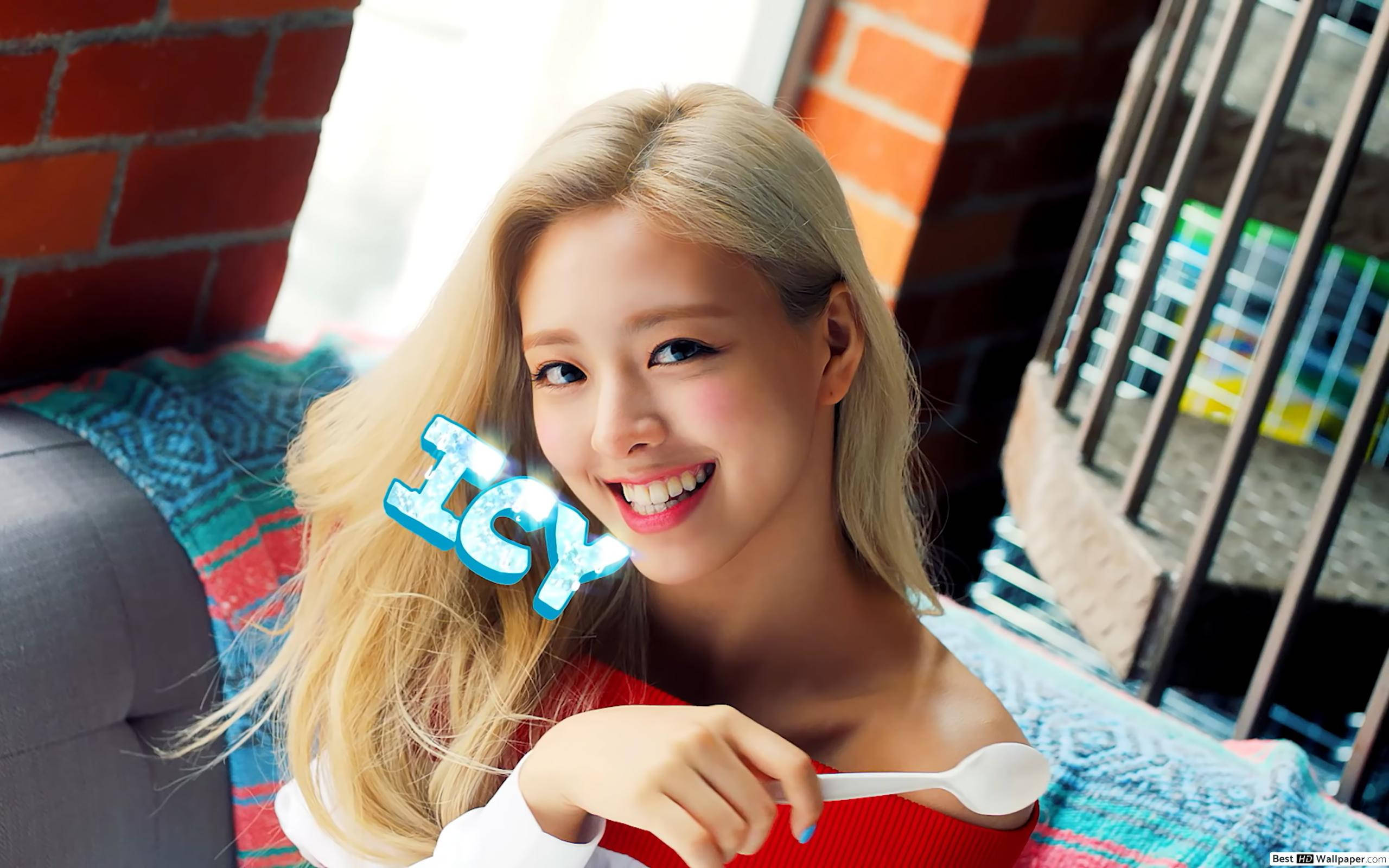 ITZY 申有娜《Icy》 壁紙 — 一位微笑的金髮女子拿著湯匙，在磚牆和彩色毯子前展示一個冰冷的圖案。