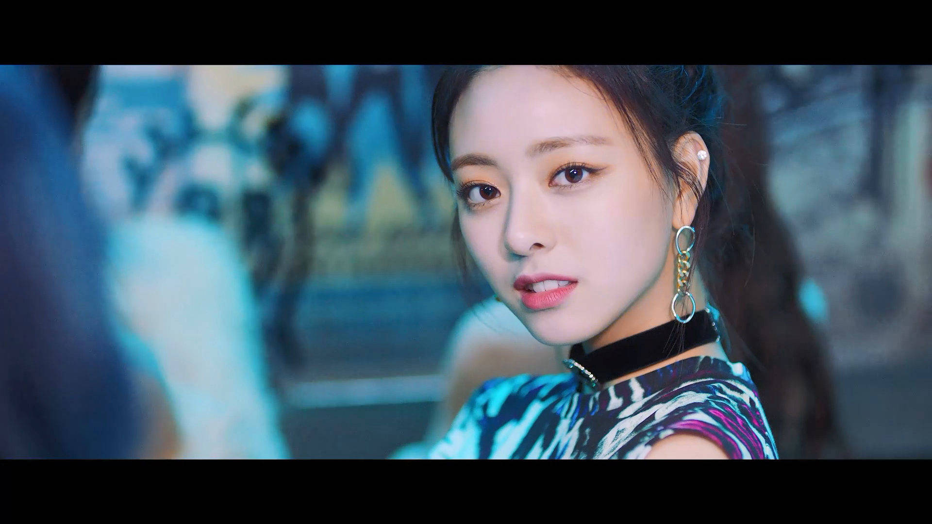 ITZY 申有娜《Wannabe》 壁紙 — 一位深色頭髮、妝容大膽的年輕女性在冷藍色燈光下直視鏡頭，身穿圖案上衣和大圈耳環。