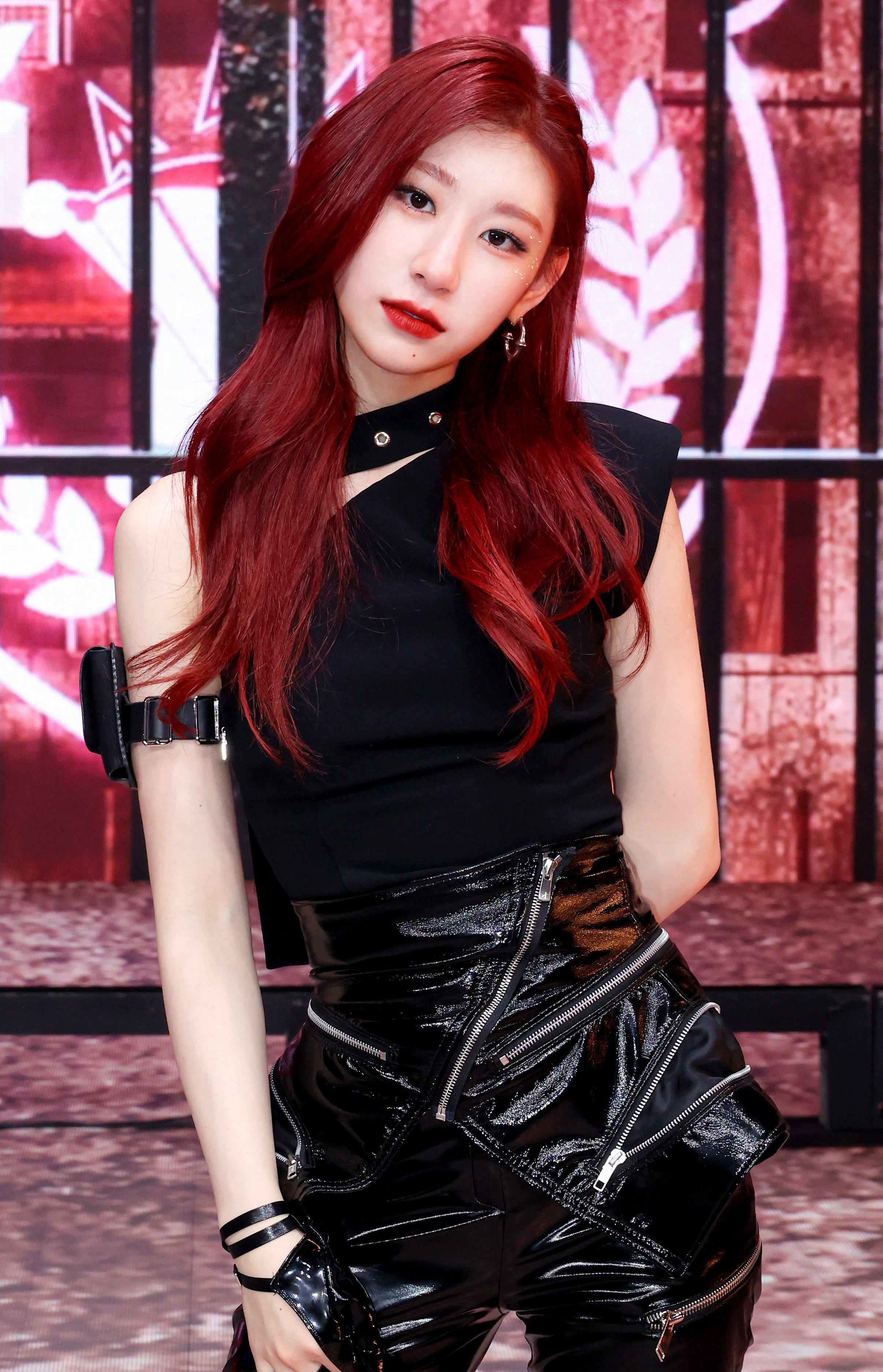 ITZY 申留真《Mafia In The Morning》 壁紙 — 一位擁有鮮豔紅髮的女性，穿著帶有拉鍊和帶子的黑色服裝，背景是發光的紅白圖案。