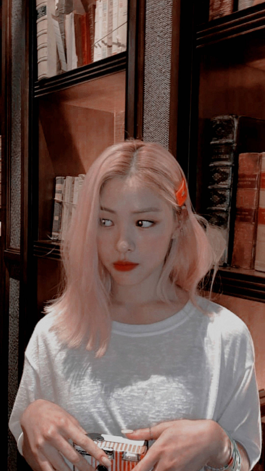 ITZY 申留真 自拍 壁紙 — 一位粉紅色頭髮、紅唇的年輕女性在裝滿書本的深色木書架前拿著一個盒子。