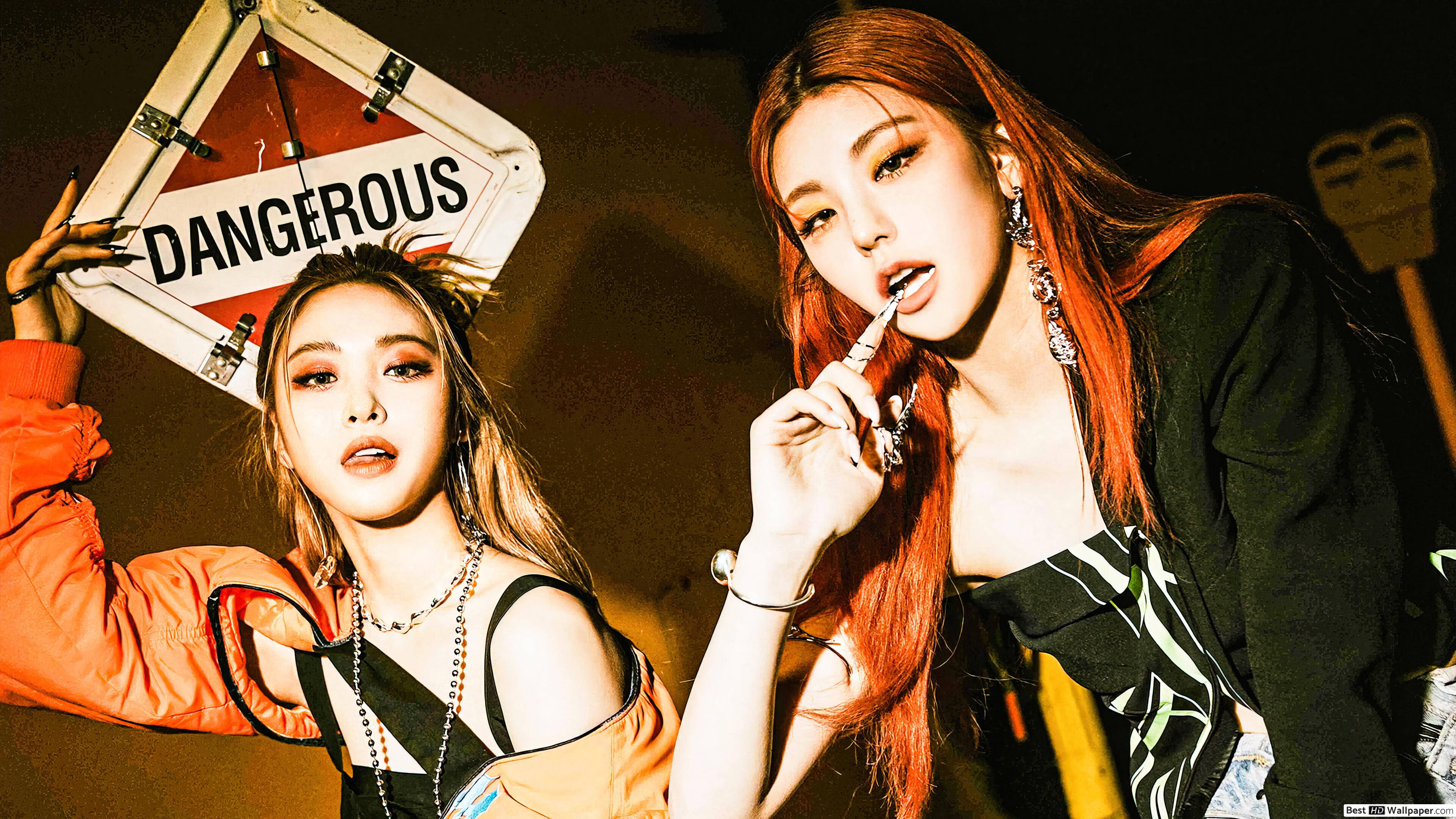 ITZY 申留真與黃藝智 壁紙 — 兩位時尚女性在夜晚戲劇性地擺姿勢，一人手持「DANGEROUS」標誌，另一人在溫暖燈光下舔唇膏管。