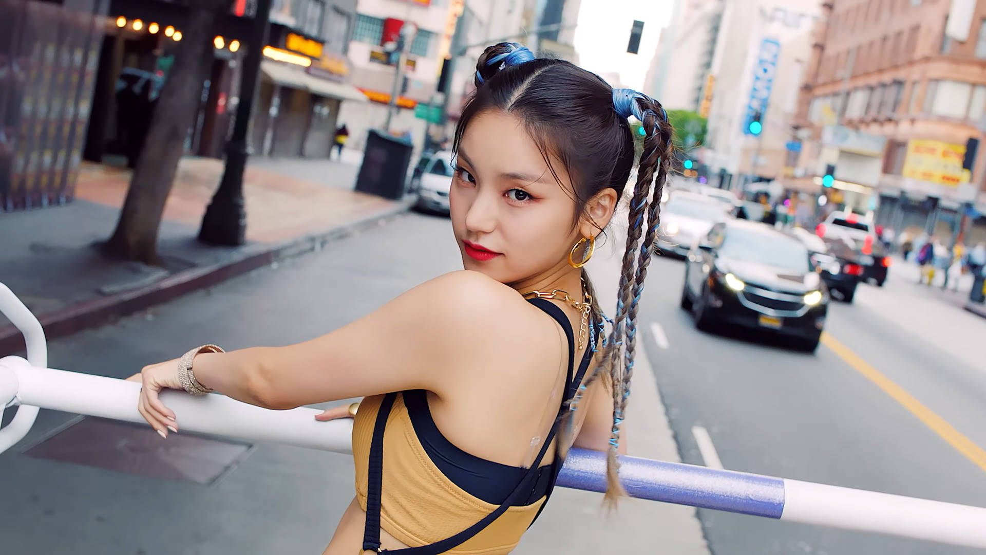 ITZY 隊長黃藝智 壁紙 — 一位年輕女子，辮子末端染藍，倚靠在繁華城市景觀的街道欄杆上，回頭凝視，紅唇大膽，戴著金色耳環。