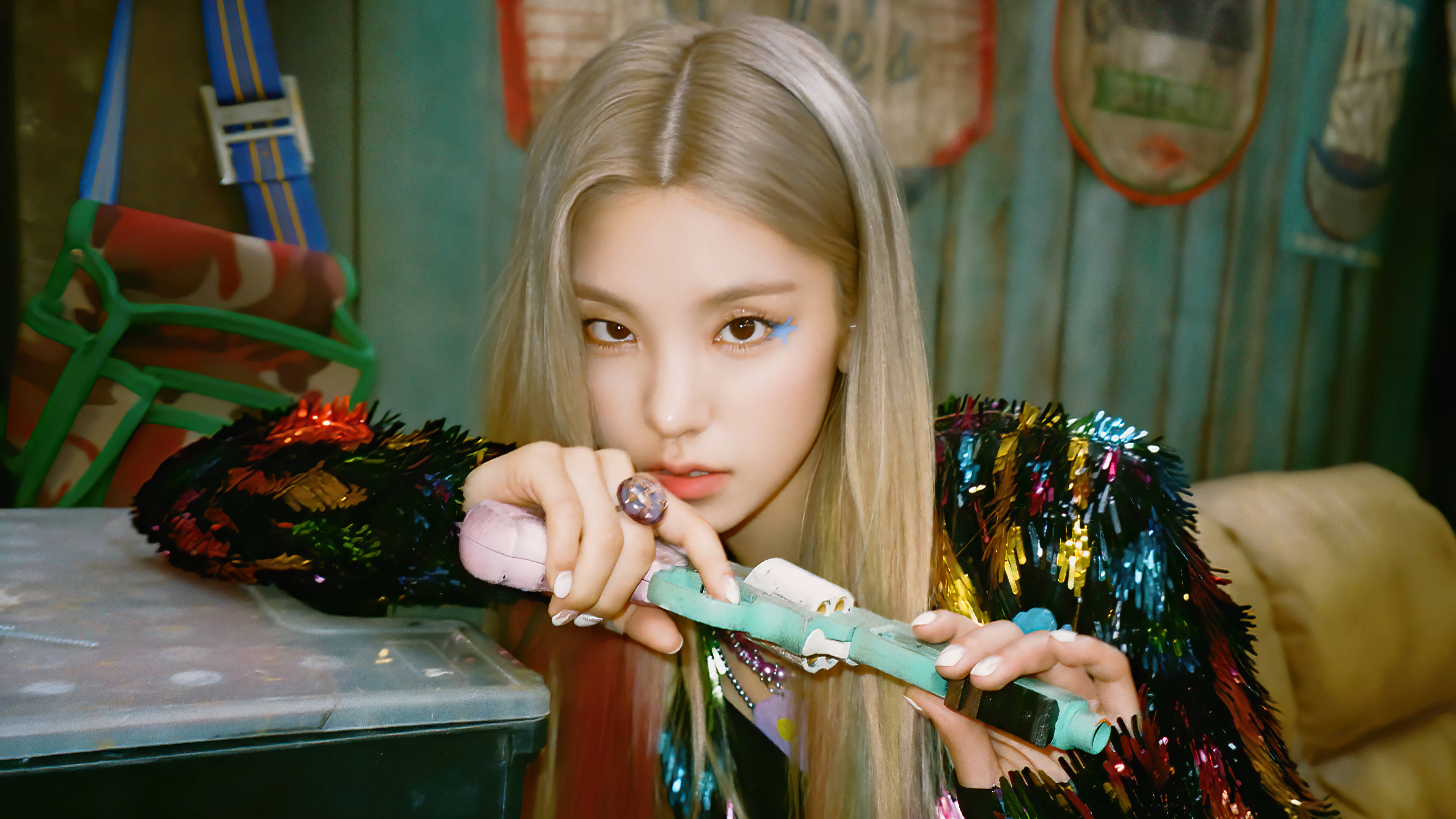 ITZY 黃藝智《Not Shy》 壁紙 — 一位金髮碧眼的年輕女子，化著大膽的藍色眼妝，穿著閃亮的金屬絲夾克，手持彩色玩具槍。