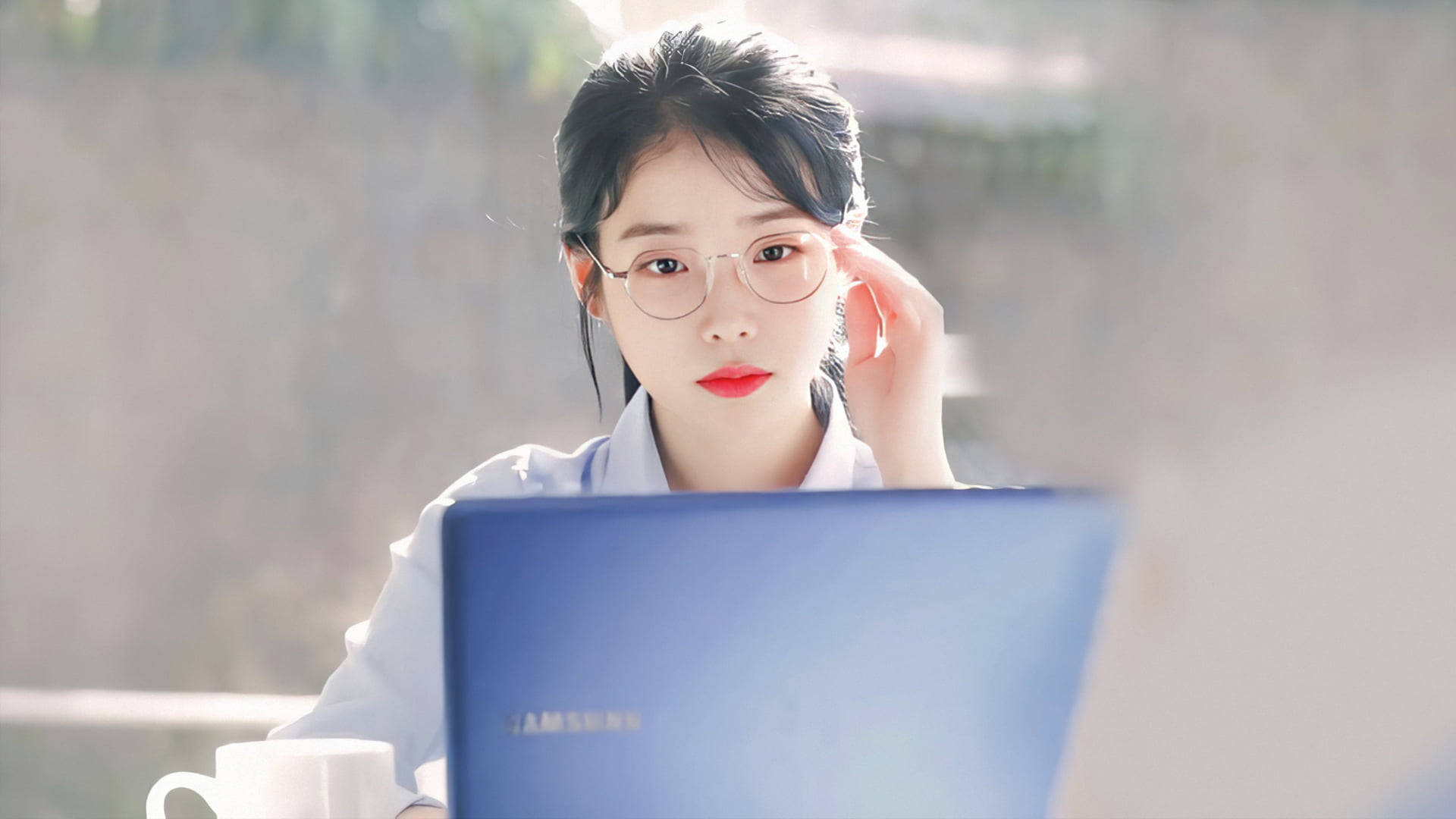 IU 配戴透明眼鏡 壁紙 — 一位戴眼鏡、紅唇的年輕女性專注地看著她的手提電腦，陽光從她身後的窗戶照射進來。