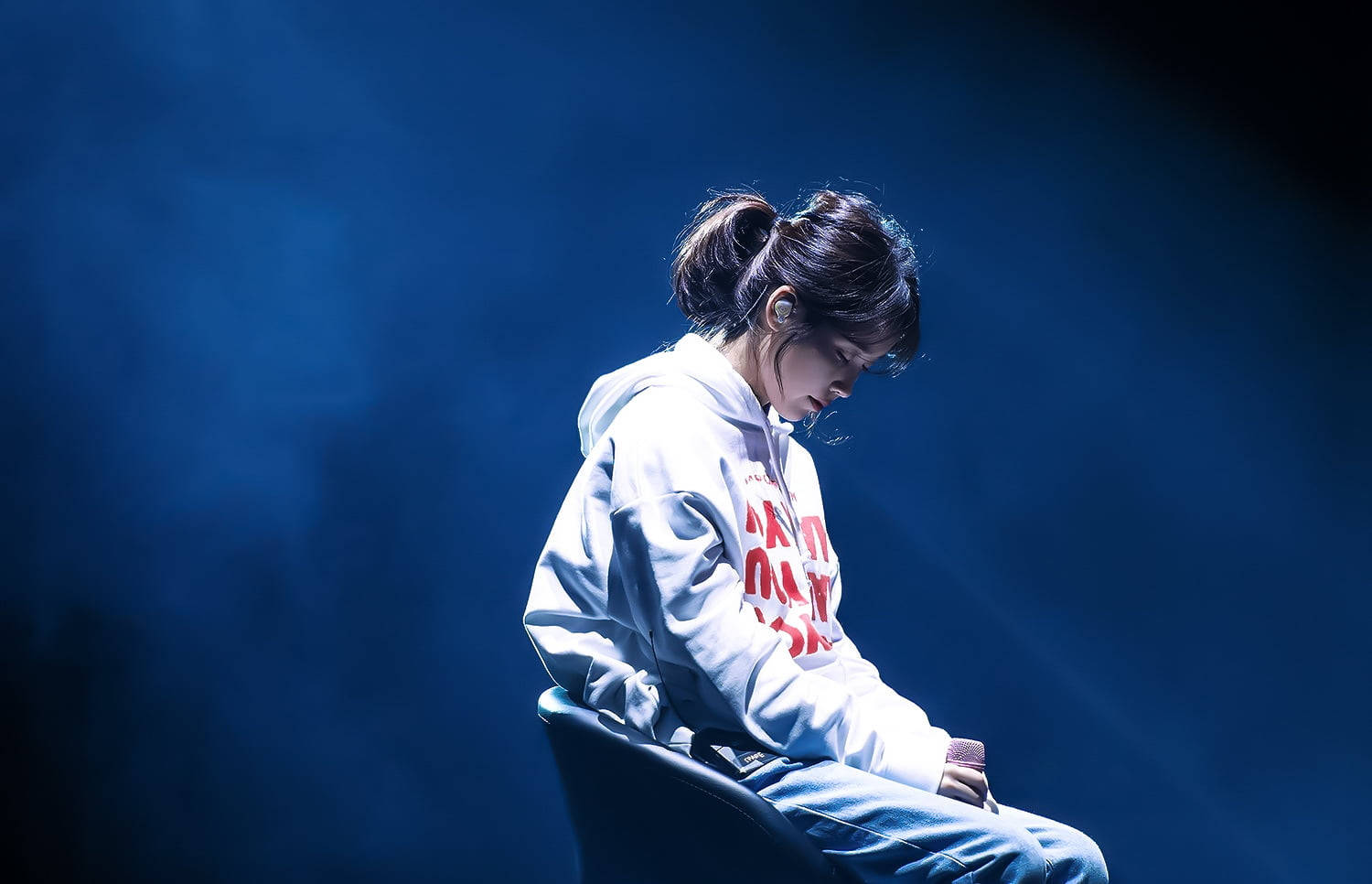 IU在深藍背景前點頭 壁紙 — 一位身穿白色連帽衫的年輕女性獨自坐在椅子上，沐浴在戲劇性的藍色舞台燈光中，煙霧在她周圍繚繞。