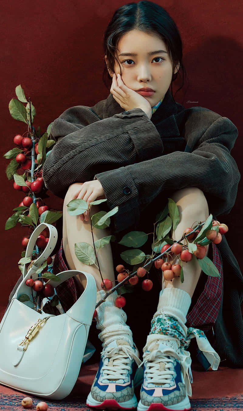 IU為《Elle Men China》拍攝時尚寫真 壁紙 — 一位身穿燈芯絨外套的年輕女子，在深紅色背景前，若有所思地坐在一個白色手袋和櫻花枝旁。