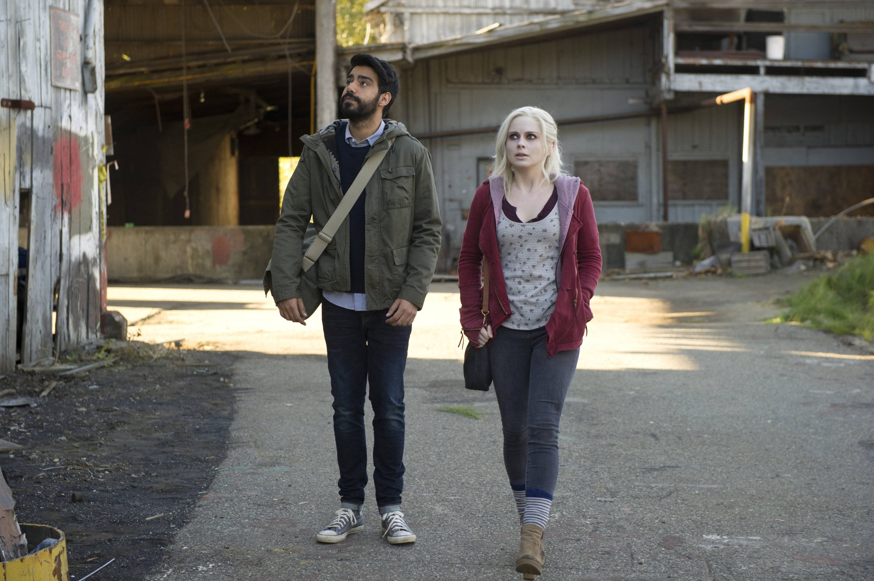《iZombie》莉芙·摩爾與醫生拉維·查克拉巴蒂 壁紙 — 兩個人沿著一條陽光照耀的碎石路行走，旁邊是一座破舊的工業建築，油漆剝落，金屬生鏽。