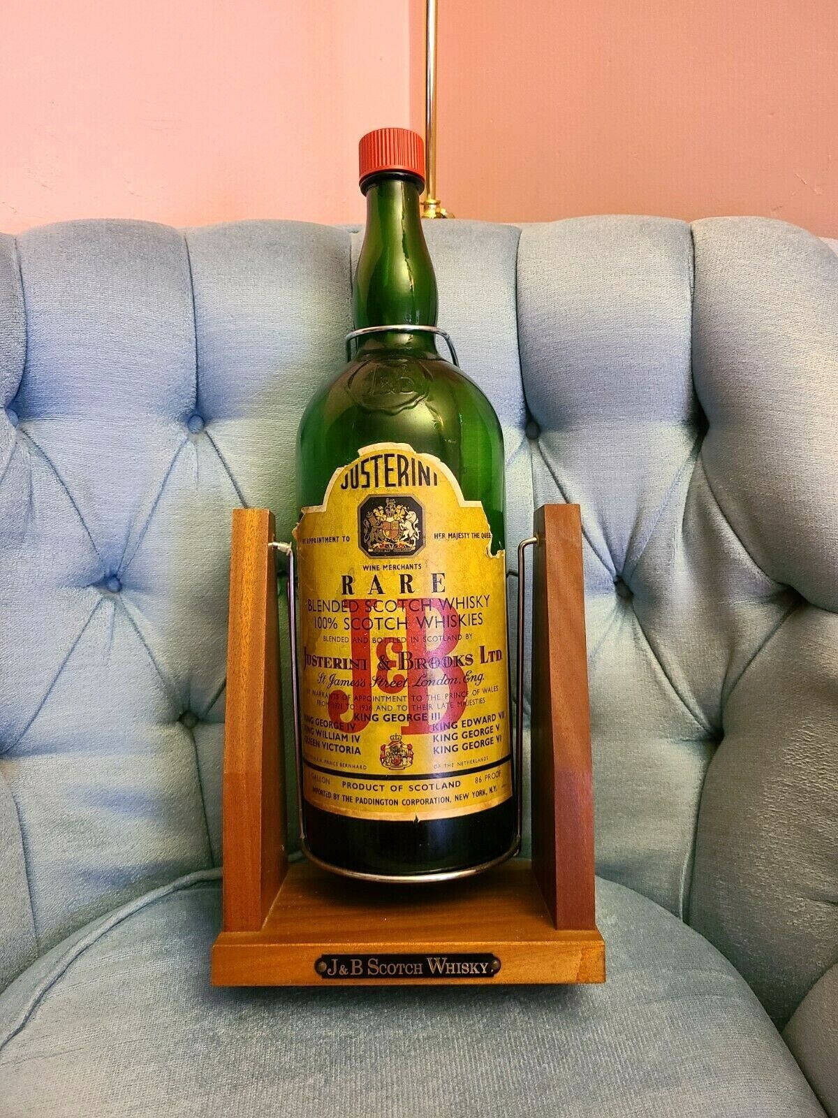J&B Rare 威士忌巨型瓶 壁紙 — 一瓶復古綠色Justerini & Brooks稀有調和蘇格蘭威士忌，優雅地放置在淺藍色簇絨沙發上的木架上，背景是柔和的粉紅色牆壁。