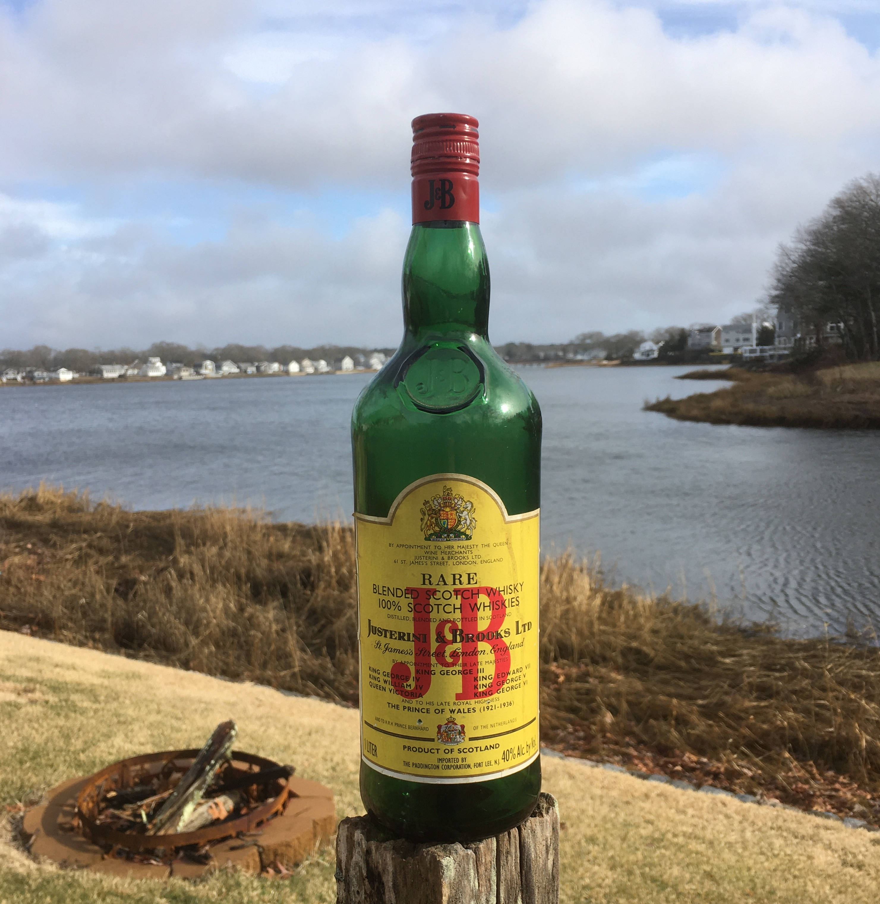 J&B Rare 蘇格蘭威士忌 – 純粹封存於瓶中 壁紙 — 一瓶罕見的蘇格蘭威士忌綠色瓶子，立在多雲天空下平靜水邊的木樁上。
