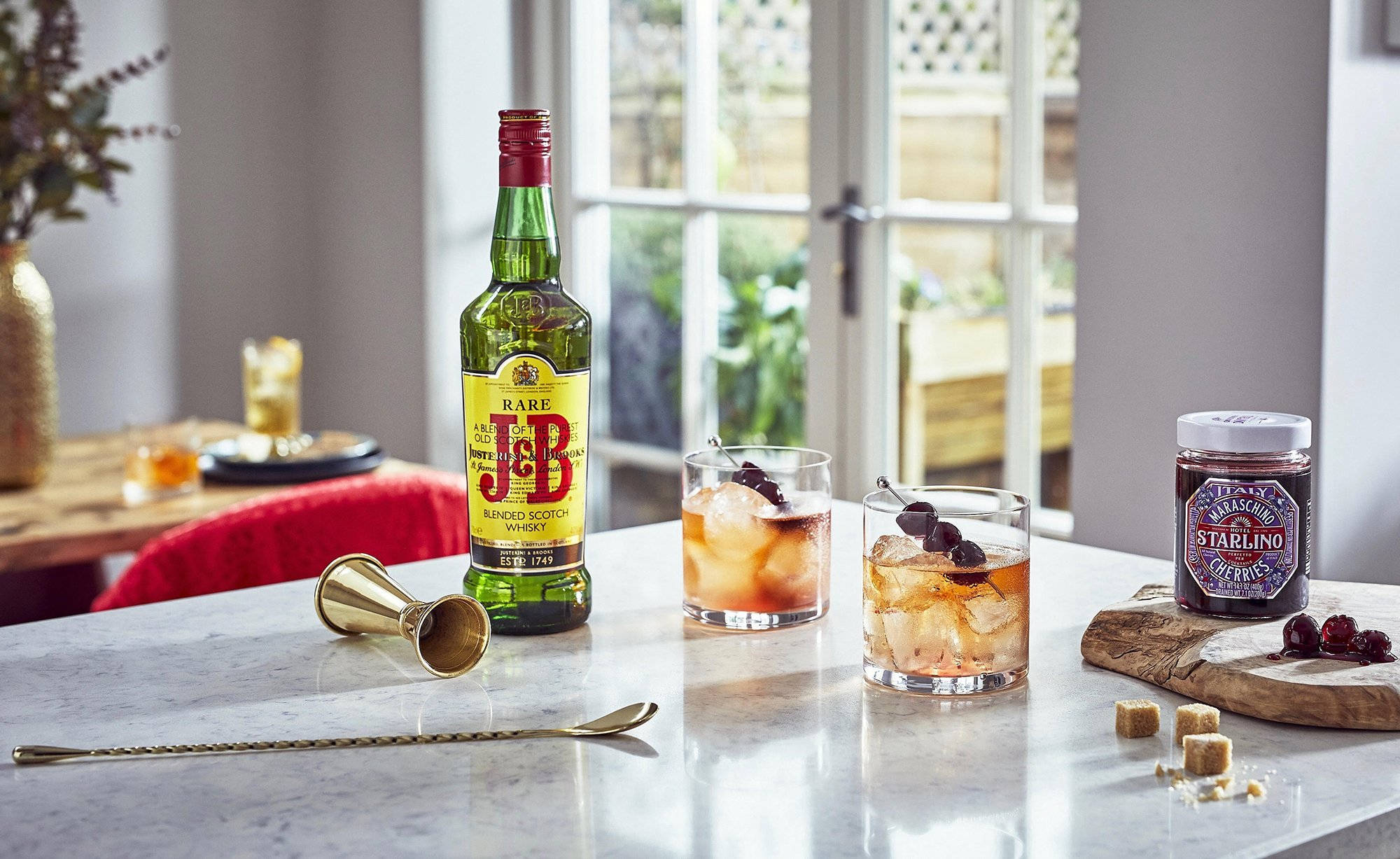 J&B 威士忌與飲品 壁紙 — 光滑的大理石枱面上展示了一瓶J&B威士忌、兩杯加冰和櫻桃的雞尾酒、一罐Starling果醬以及酒吧工具，沐浴在柔和自然光中。
