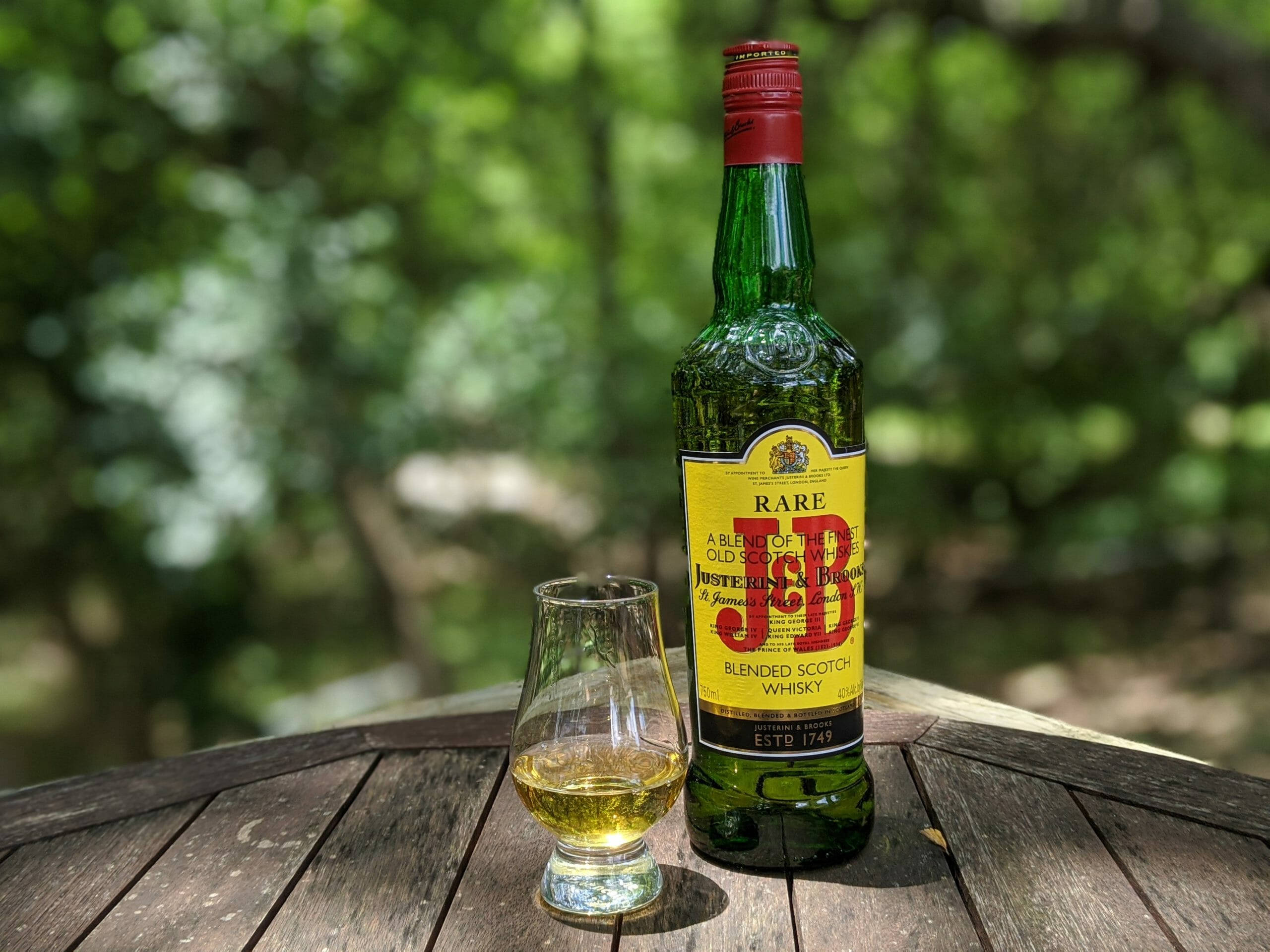 J&B蘇格蘭威士忌酒杯 壁紙 — 一瓶J&B Rare威士忌和一個裝滿的品酒杯放在戶外一張風化的木桌上，背景是柔焦的茂密綠葉。