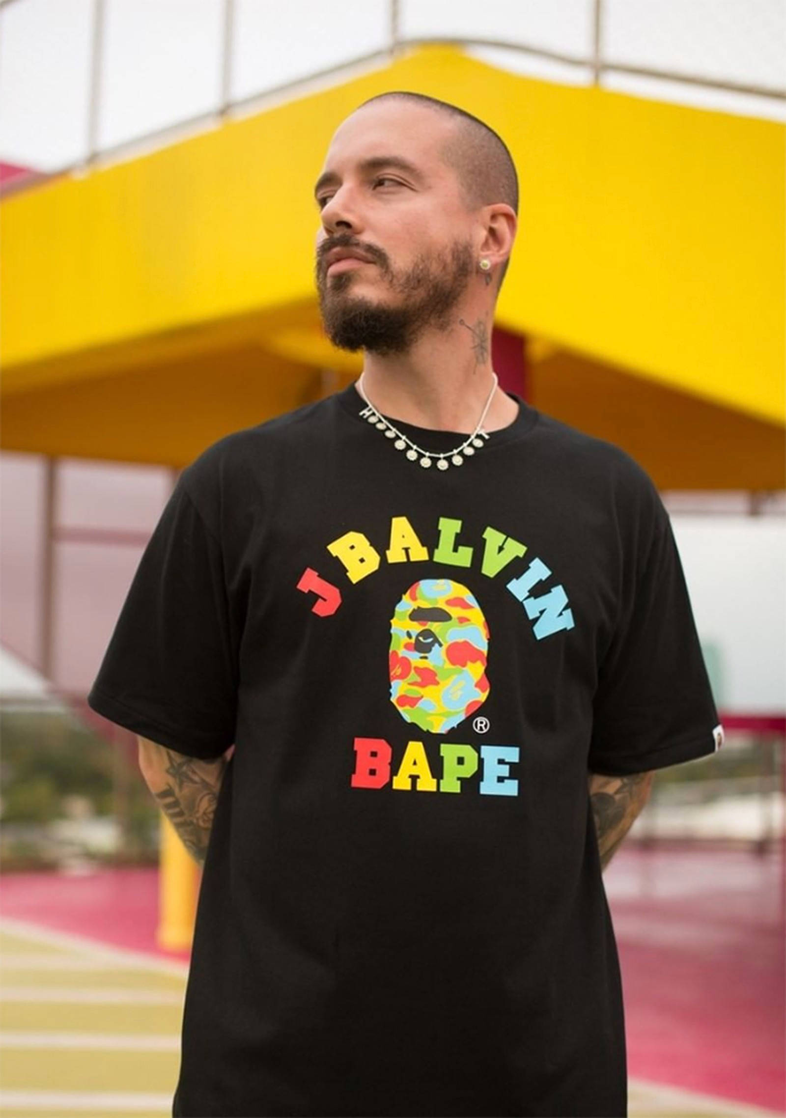 J Balvin A Bathing Ape聯名款 壁紙 — 一個留鬍鬚和有紋身的男人，穿著一件黑色T恤，上面有色彩繽紛的Bape標誌，背景是明亮的黃色建築。