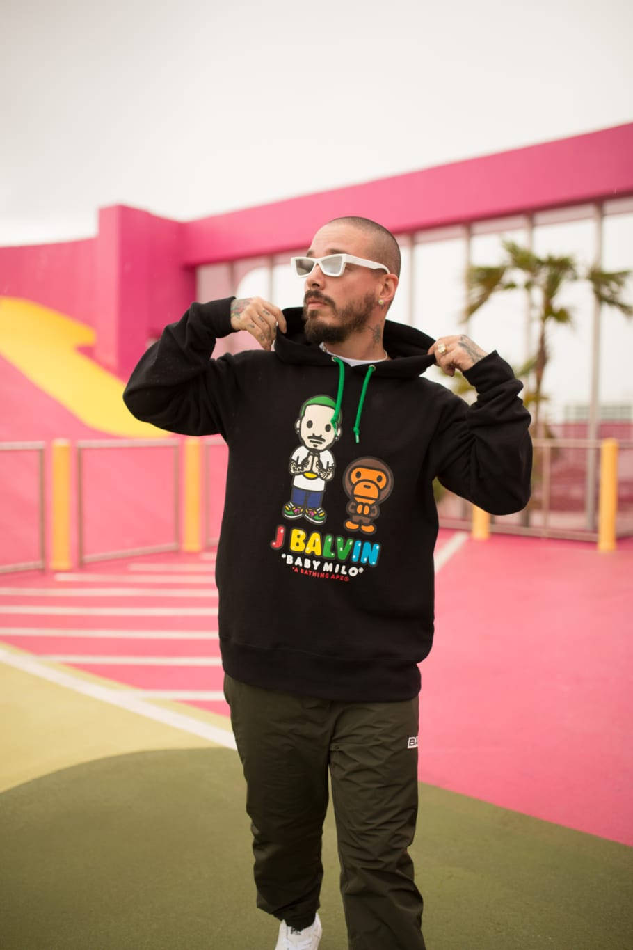 J Balvin Baby Milo聯名款 壁紙 — 一個穿著帶有卡通圖案黑色連帽衫的男人拉起帽子，站在色彩繽紛的粉紅色和黃色球場上。