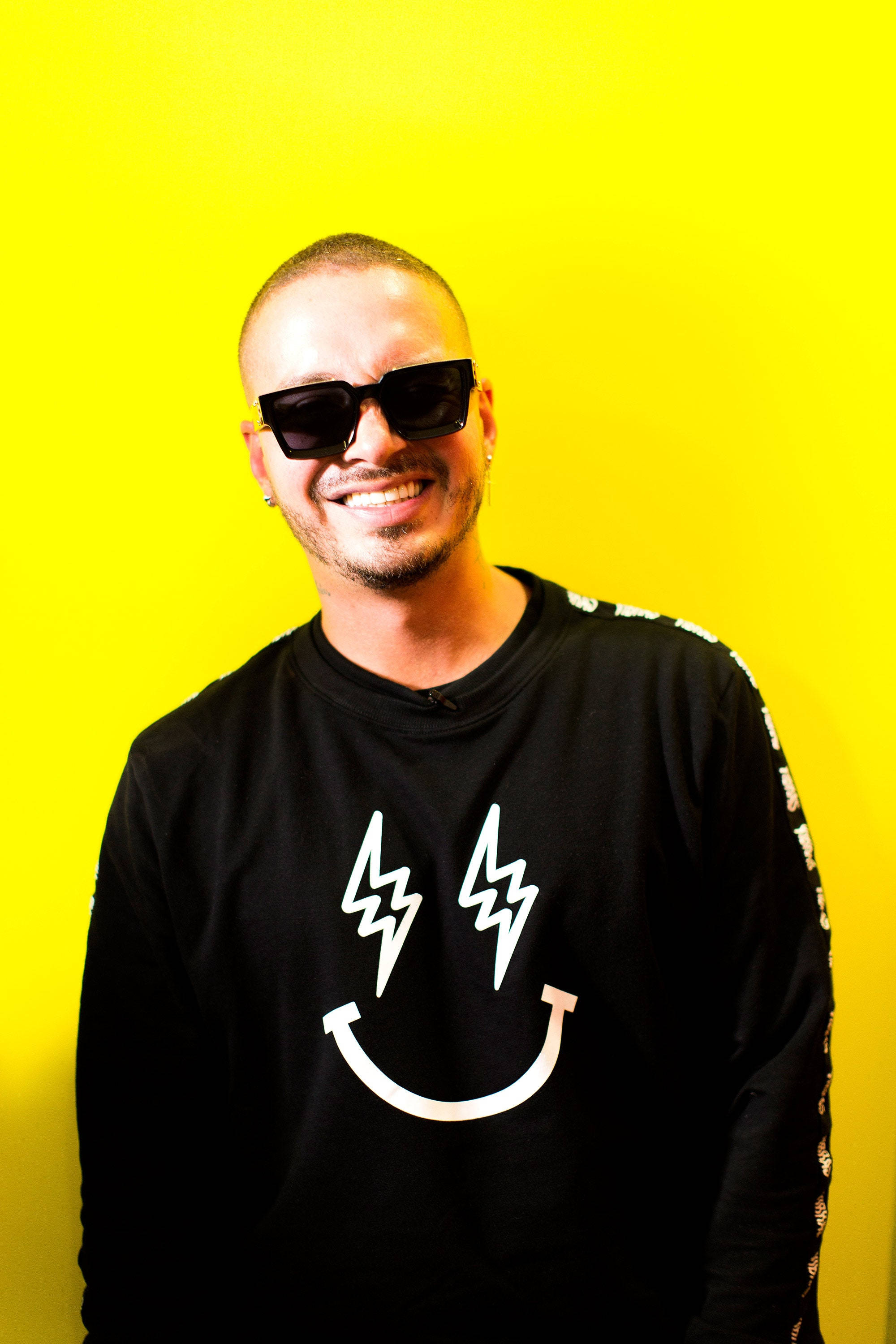 J Balvin GEF 聯名衛衣 壁紙 — 一個微笑的男人，戴著黑色太陽眼鏡，身穿印有白色閃電笑臉的黑色襯衫，站在鮮黃色背景前。