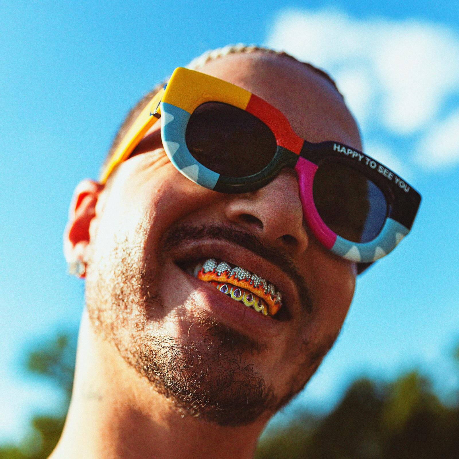 J Balvin JOSE 專輯封面 壁紙 — 一個微笑的男人戴著彩色太陽眼鏡和鑲有寶石的牙套，在明亮的藍天下。