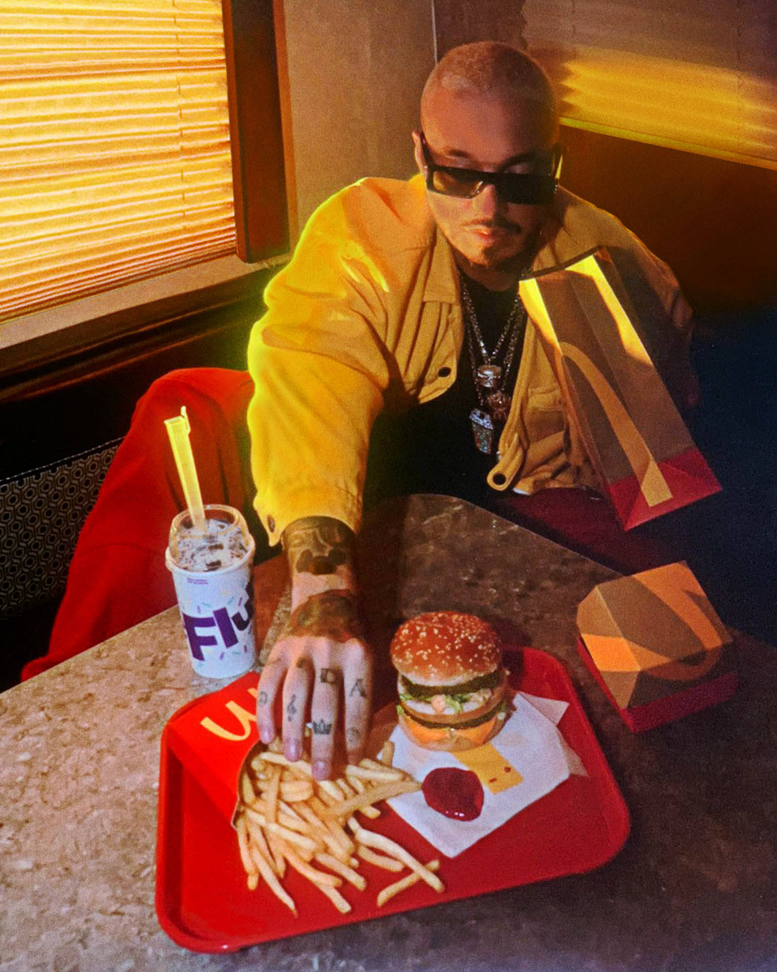 J Balvin 吃漢堡薯條 壁紙 — 一名身穿黃色外套和太陽眼鏡的男子坐在餐廳餐桌旁，伸手去拿紅色托盤上漢堡和飲料旁邊的薯條。