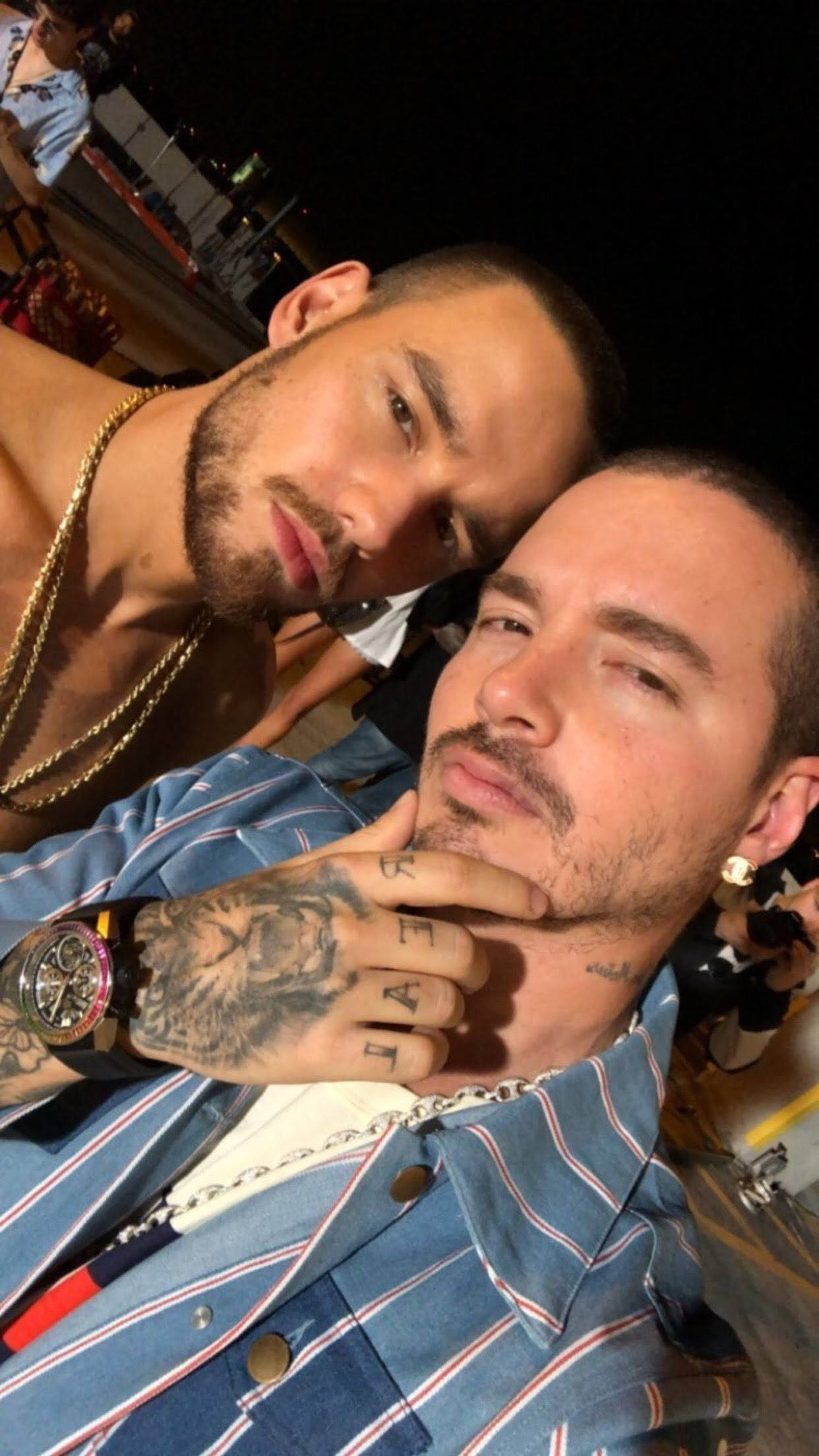 J Balvin 與 Liam Payne 壁紙 — 兩個男人在昏暗的夜晚環境中緊密擺姿勢，一個赤裸上身戴金鏈，另一個穿條紋衫，紋身的手放在下巴上。