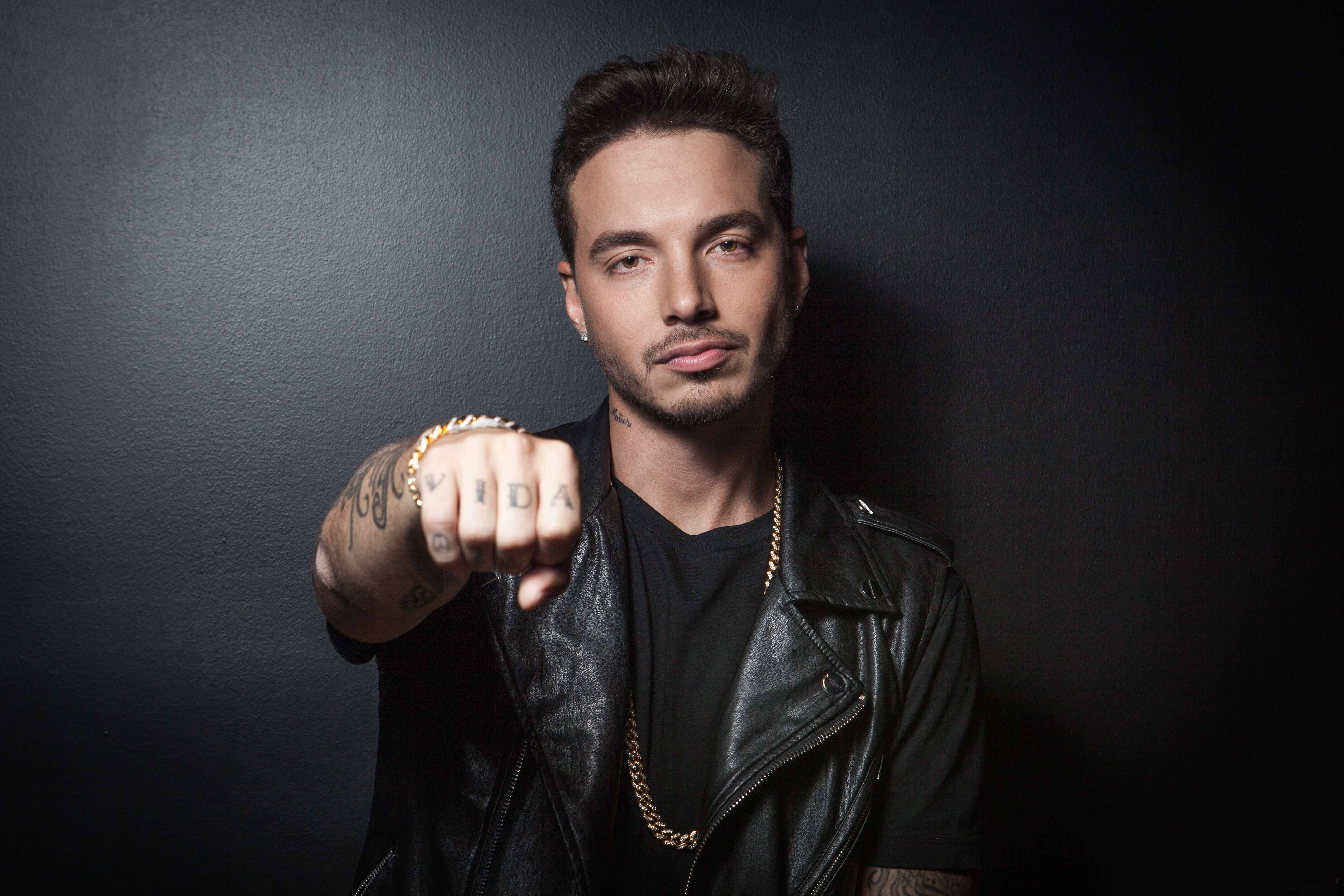 J Balvin VIDA拳頭紋身 壁紙 — 一名身穿黑色皮褸的男子，在深色紋理牆壁前，將紋身拳頭指向鏡頭。