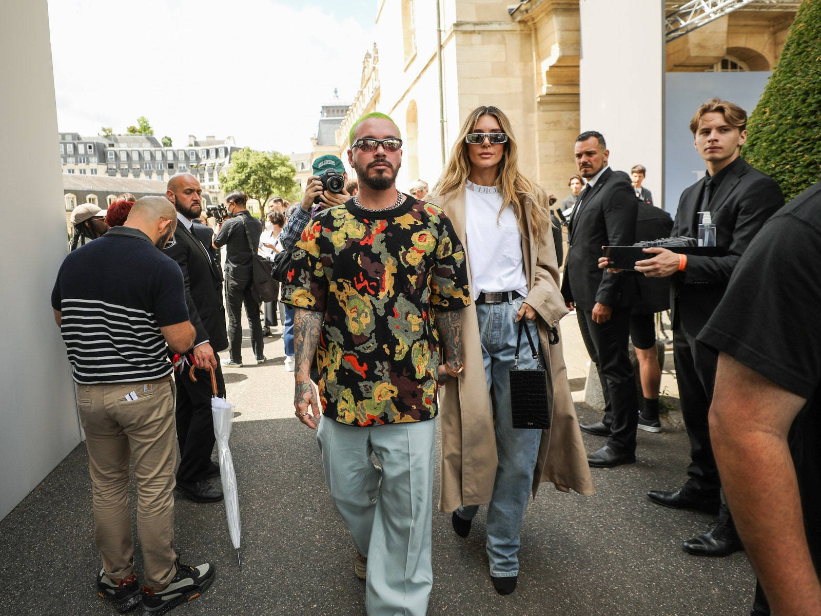 J Balvin 出席 Christian Dior 男裝秀 壁紙 — 一對時尚情侶自信地走過一個陽光普照的戶外時裝活動，周圍有攝影師和觀眾。