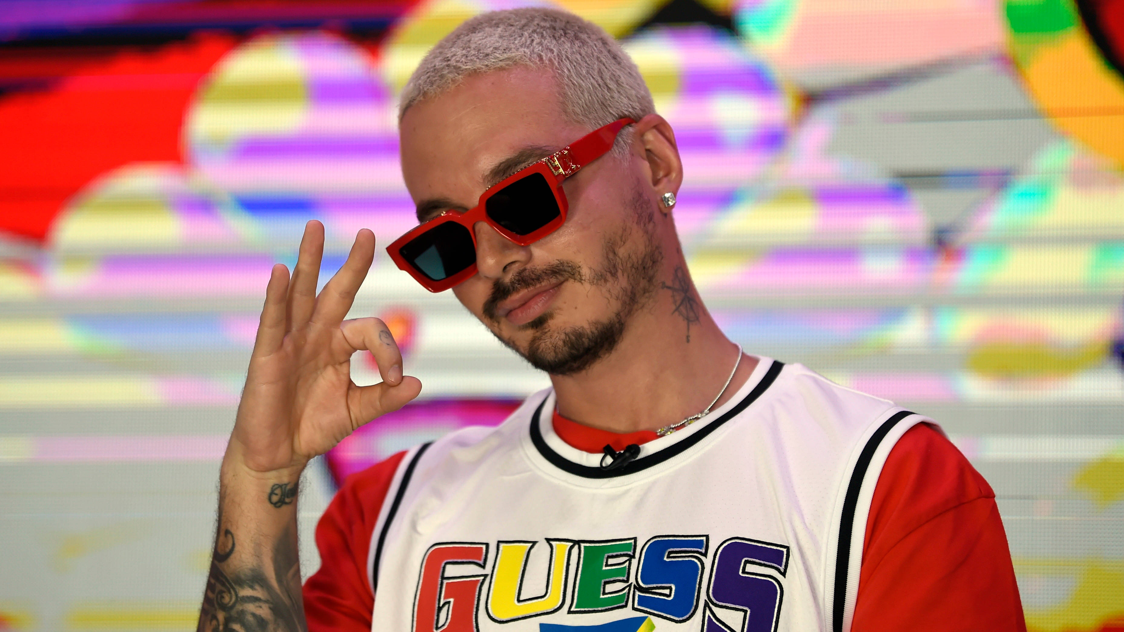 J Balvin 彩虹 Guess 衣著 壁紙 — 一個頭髮漂白、戴紅色太陽眼鏡的男人，穿著色彩繽紛的Guess球衣，在充滿活力的抽象背景前做出OK手勢。