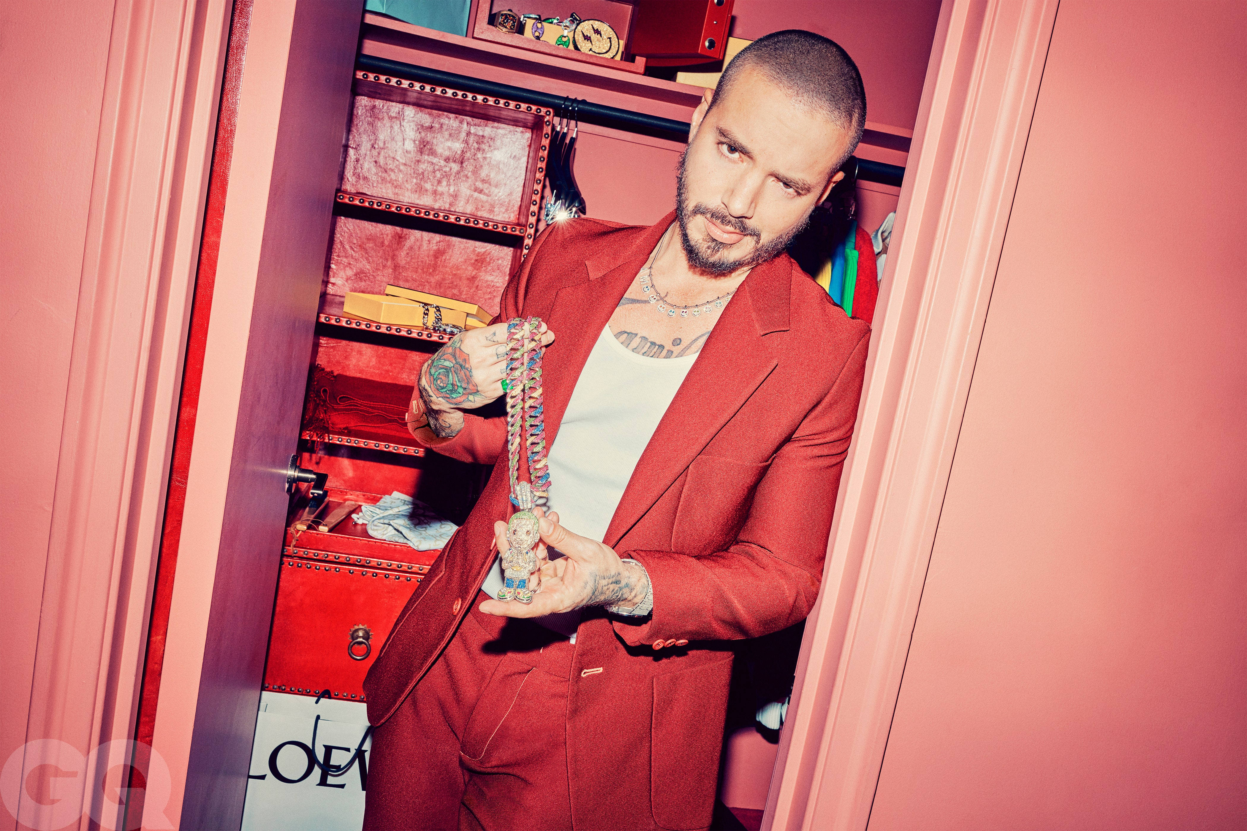 J Balvin 為 GQ 拍攝 壁紙 — 一個穿著紅色西裝的男人站在粉紅色衣櫃裡，手持珠鍊，手臂上可見紋身。