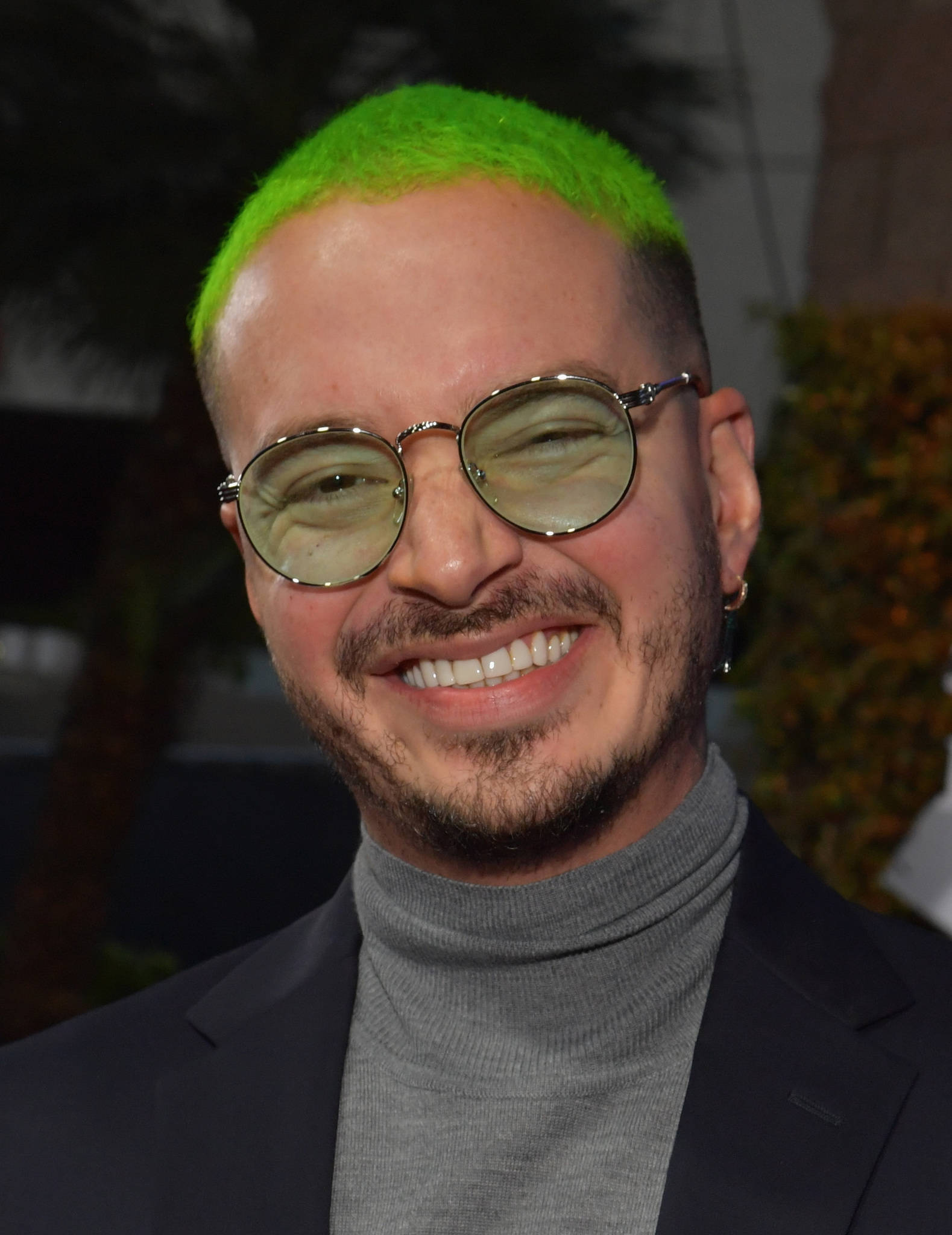 J Balvin 綠髮造型 壁紙 — 一位擁有亮綠色頭髮和圓框眼鏡的男子，身穿深色西裝和灰色高領毛衣，溫暖地微笑。