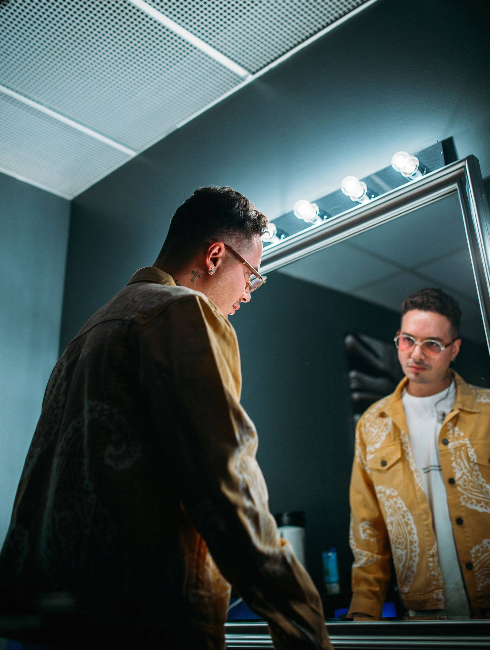 J Balvin 鏡中倒影 壁紙 — 一個穿著黃色圖案外套的男人在後台凝視著明亮鏡子中的倒影。