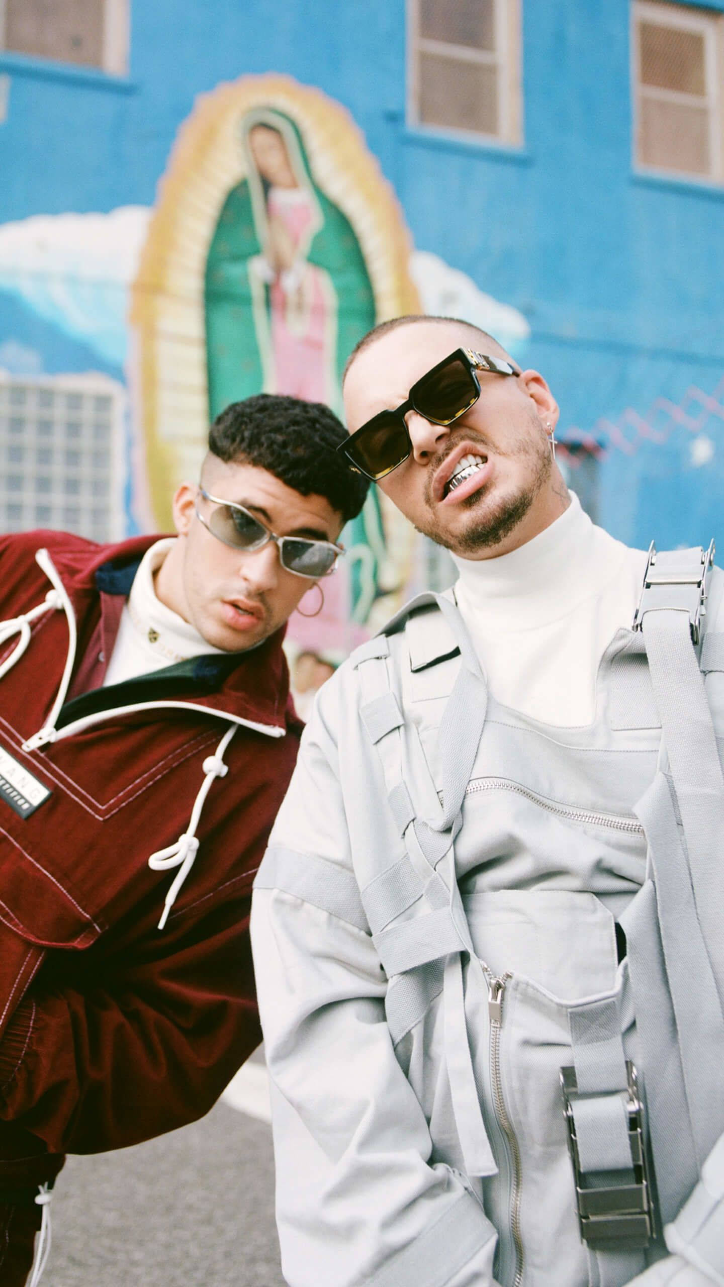 J Balvin與Bad Bunny同框 壁紙 — 兩位時尚男士戴著太陽眼鏡，自信地在充滿活力的藍色瓜達盧佩聖母壁畫前擺姿勢。
