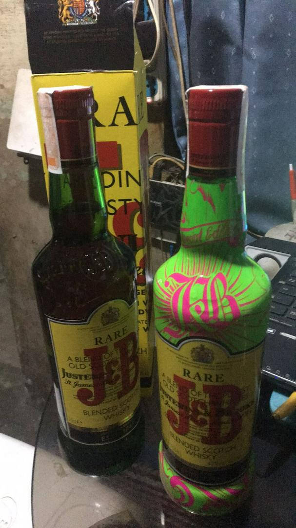 J&B瓶與J&B珍藏紋身瓶 壁紙 — 兩瓶罕見的J&B威士忌酒瓶，一瓶標準版，一瓶定制塗成綠色和粉紅色，放在玻璃桌上，旁邊有一台筆記型電腦和雜亂的背景。