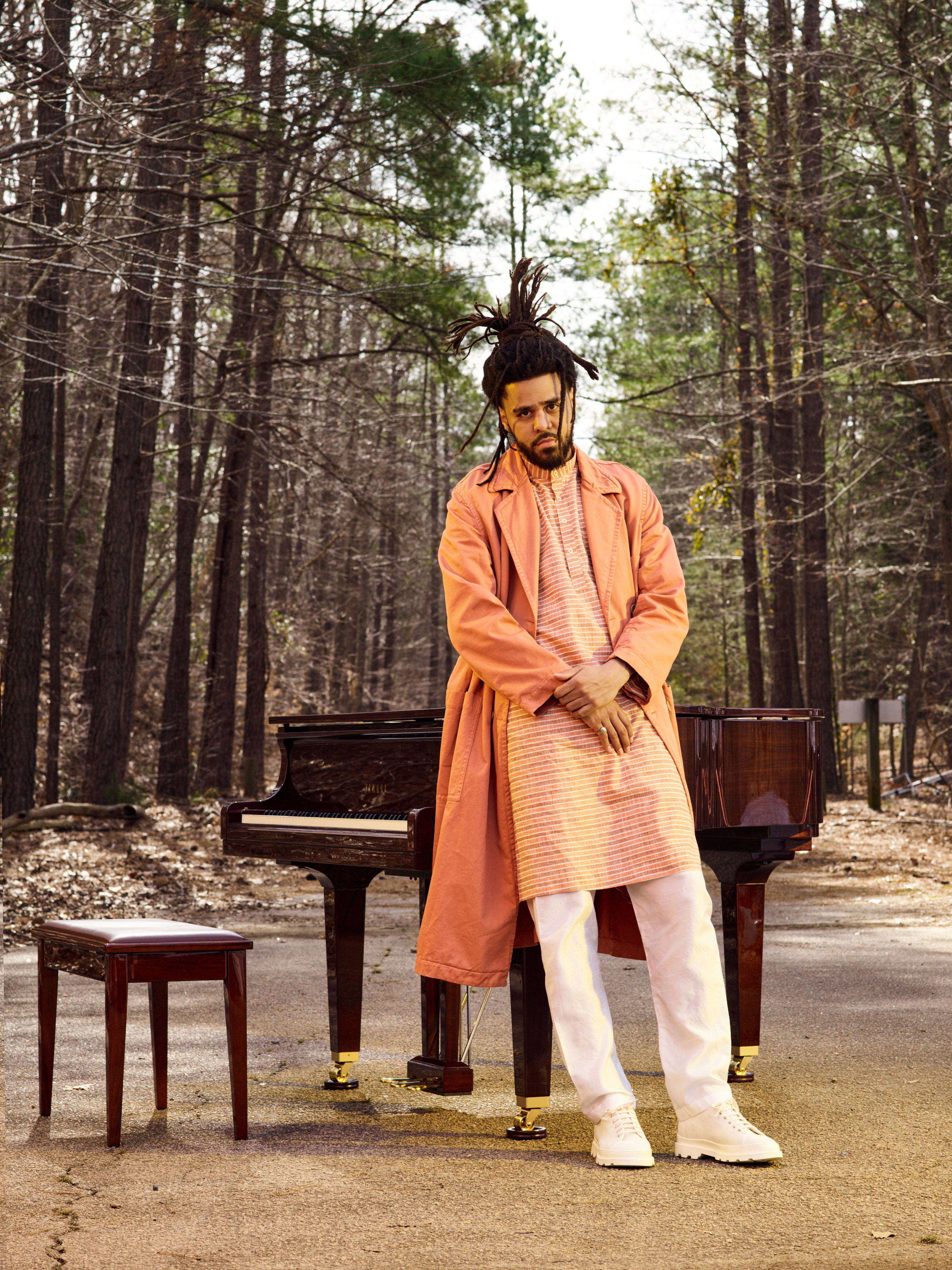 J Cole GQ雜誌封面 壁紙 — 一名身穿橙色外套的男子站在寧靜森林中的三角鋼琴旁，樹木光禿，光線柔和。