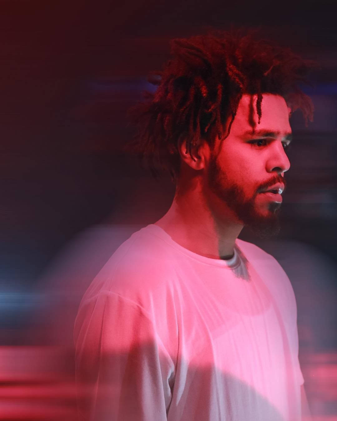 J Cole 動態效果 壁紙 — 一名留著辮子頭和鬍鬚的男子，在模糊的背景前被戲劇性的紅藍燈光照亮。