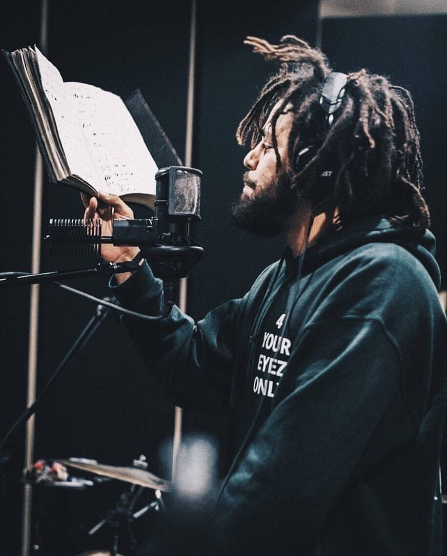 J Cole 在錄音室 壁紙 — 一位留著辮子頭、戴著耳機的音樂人在昏暗的錄音室裡拿著樂譜對著麥克風唱歌。