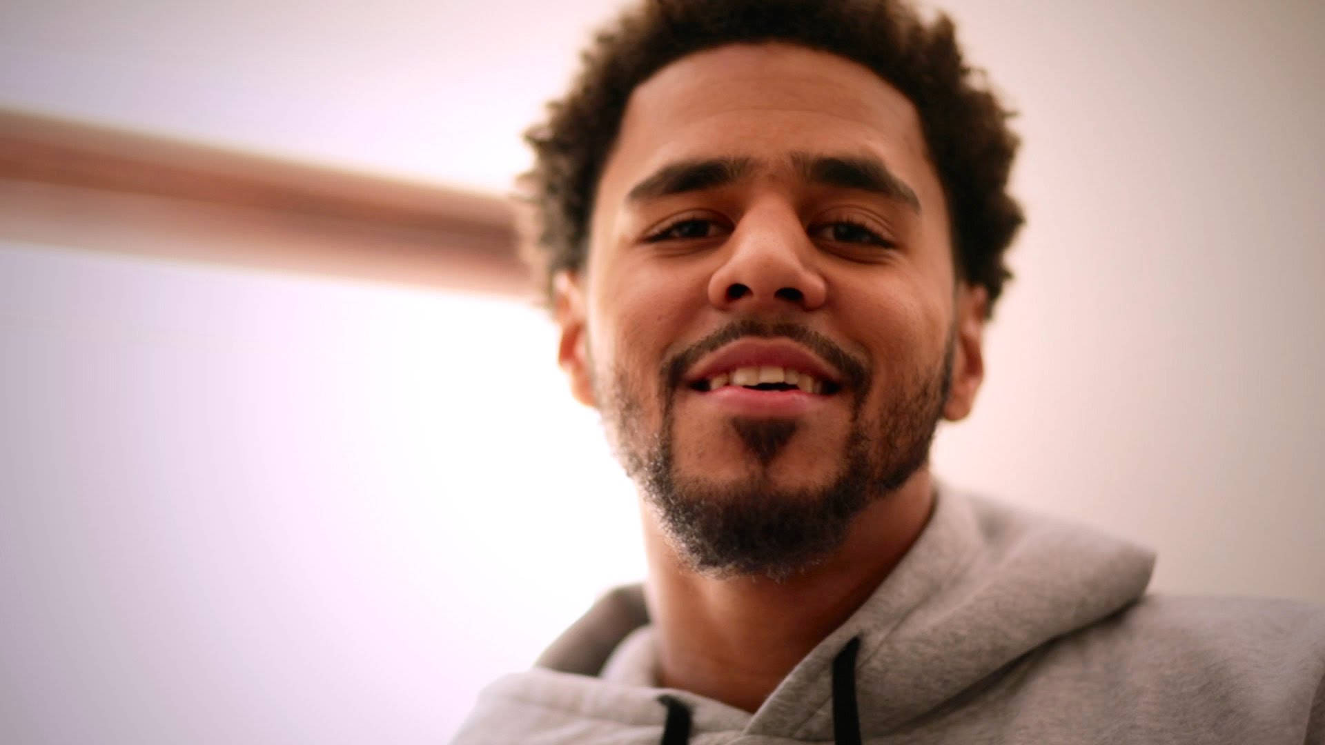 J Cole 微笑近照 壁紙 — 一個微笑的男人，有捲髮和鬍鬚，穿著灰色連帽衫，在柔和的模糊背景前的特寫肖像。