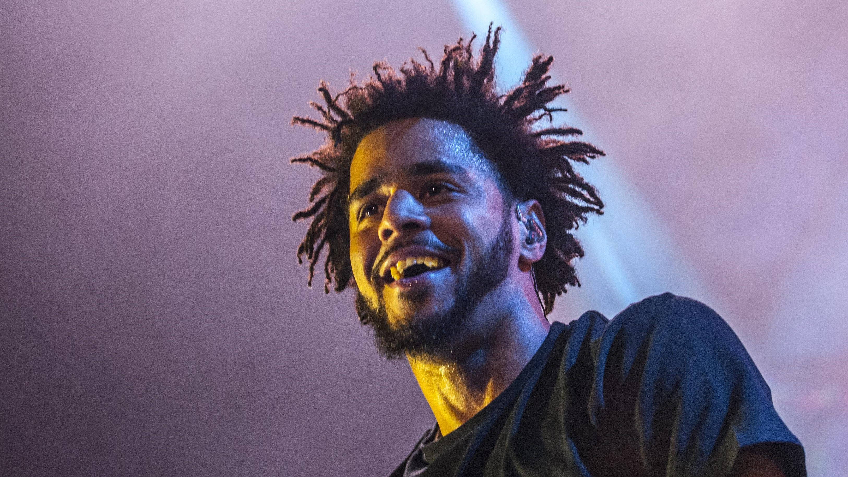 J Cole 演唱會中微笑 壁紙 — 一位面帶微笑、梳著辮子的男士在紫色和黃色的鮮豔舞台燈光下表演。