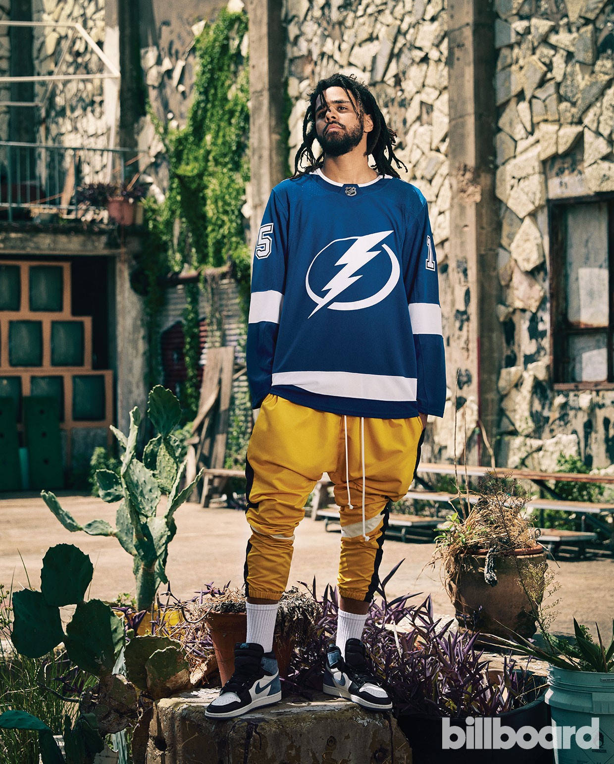 J Cole 為《Billboard》拍攝 壁紙 — 一名留著辮子頭的男子，自信地穿著藍色坦帕灣閃電隊球衣和黃色褲子，站在盆栽仙人掌和石頭建築之間。