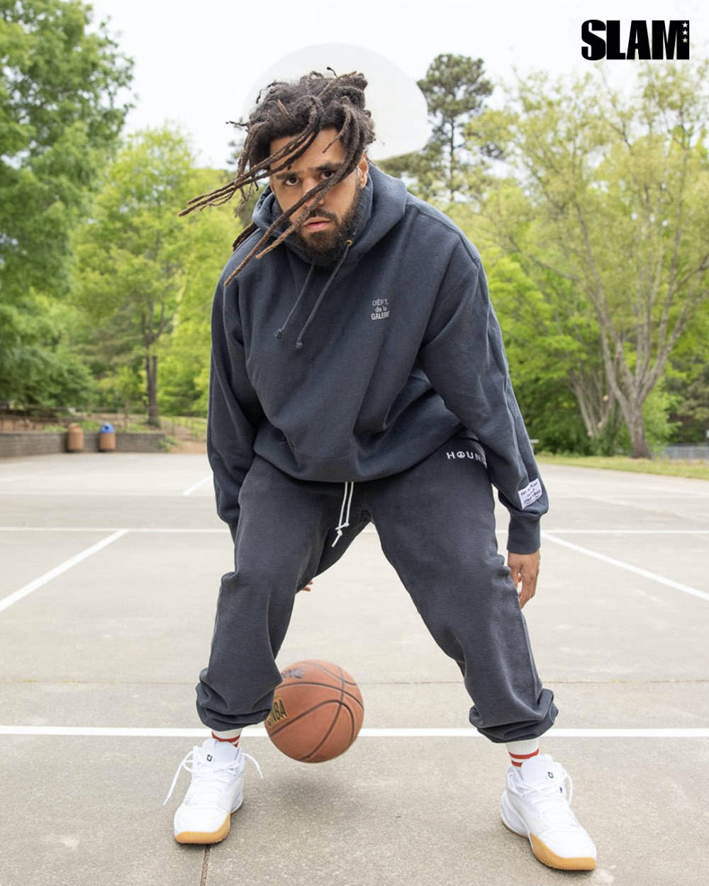 J Cole 為 SLAM 專訪 壁紙 — 一名留著辮子、身穿深色連帽衫的男子在戶外球場運球，周圍綠樹成蔭。
