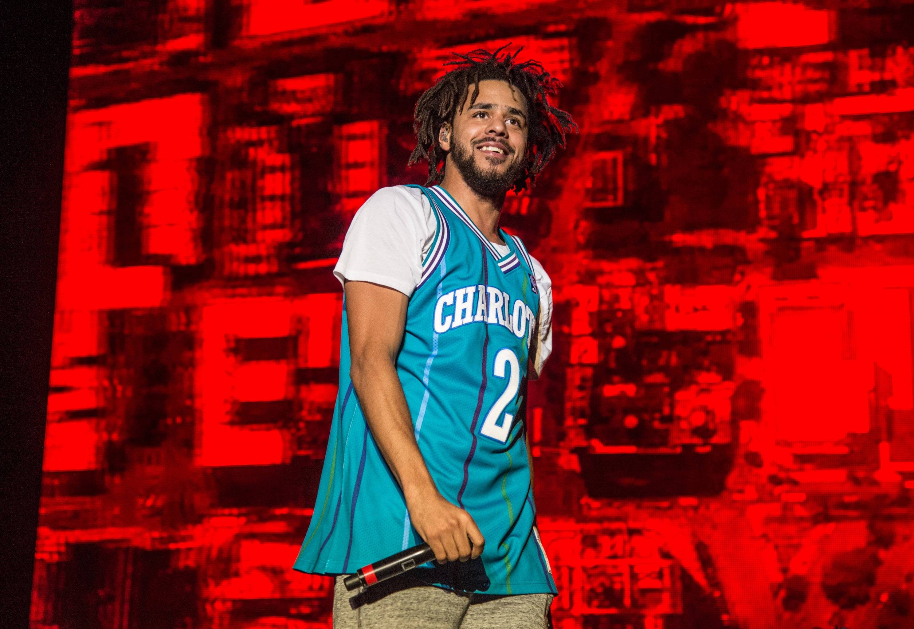 J Cole 節目穿籃球背心 壁紙 — 一個微笑的男人，梳著辮子，身穿夏洛特球衣，在充滿活力的紅色故障背景前拿著麥克風。
