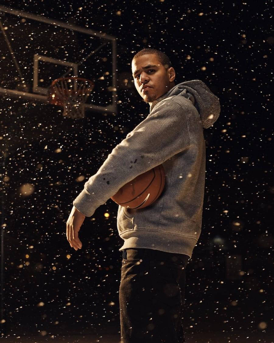 J Cole 籃球場 壁紙 — 一個穿著灰色連帽衫的年輕人在黑暗的城市環境中，背後有一個籃框，在飄雪中拿著一個籃球。