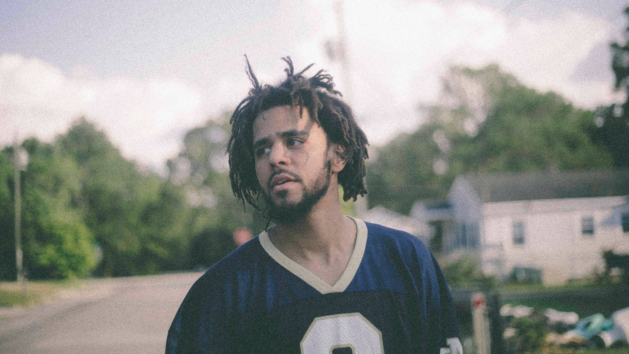 J Cole 與電影相機濾鏡 壁紙 — 一名留著辮子、身穿藍色足球球衣的年輕男子，站在多雲天空下的寧靜郊區街道上。