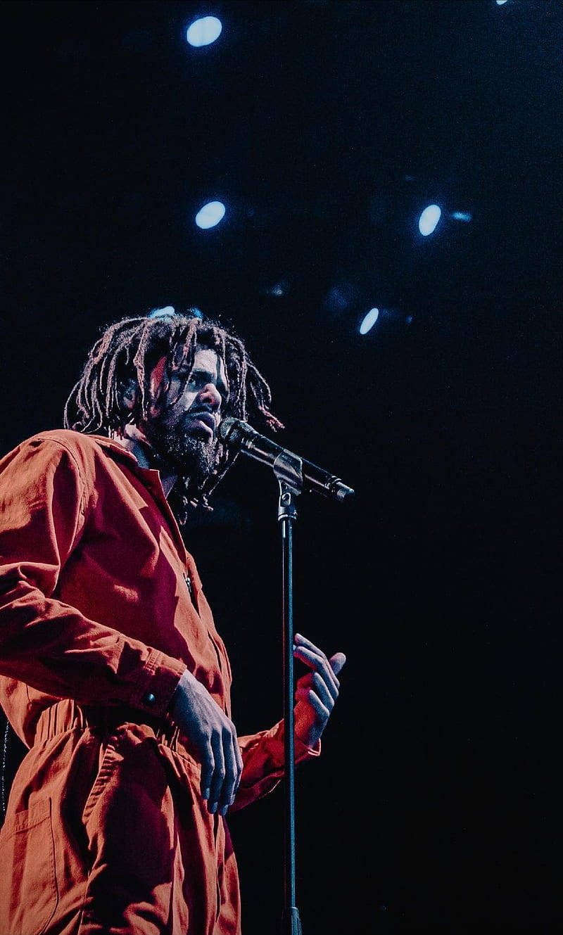 J Cole 舞台演出 壁紙 — 一位留著辮子頭的音樂家在黑暗背景下對著麥克風唱歌，舞台燈光璀璨。