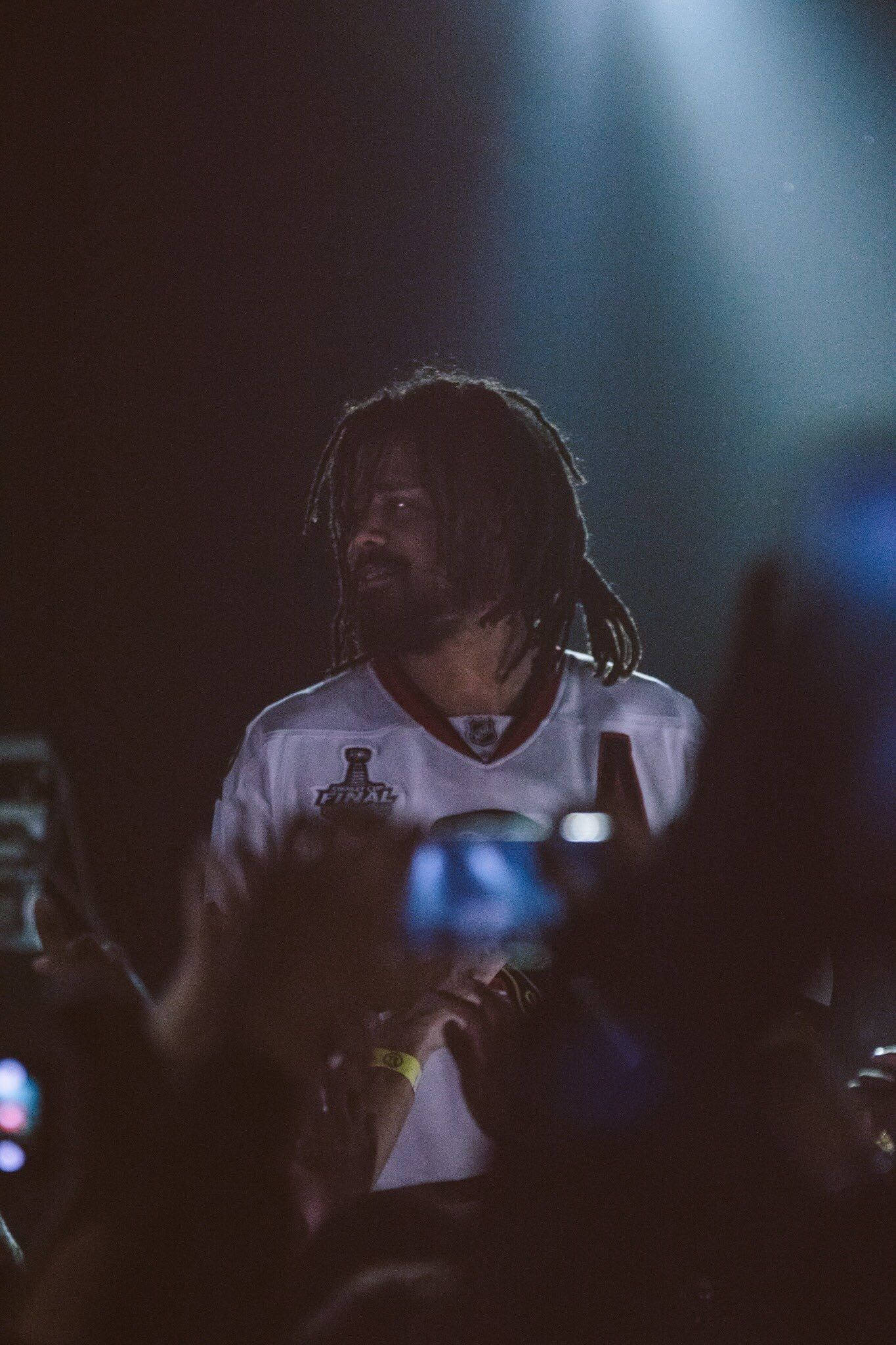 J Cole 舞台與觀眾 壁紙 — 一名留著辮子頭、身穿冰球球衣的男子，在演唱會上被聚光燈照亮，粉絲們舉著手機。