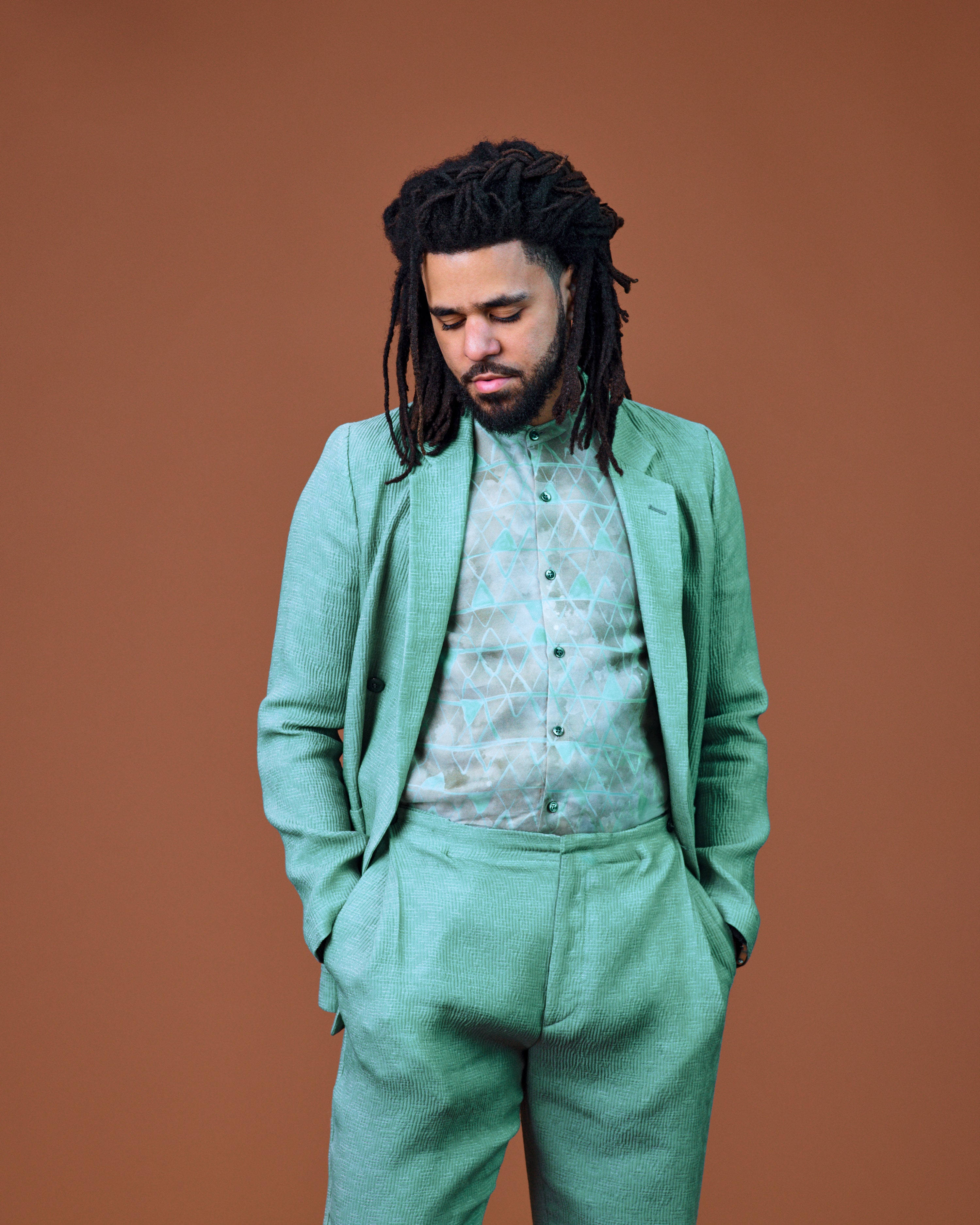 J Cole 著薄荷綠西裝 壁紙 — 一個留著辮子、身穿薄荷綠色西裝的男人，閉著眼睛站在溫暖的赤陶色背景前。