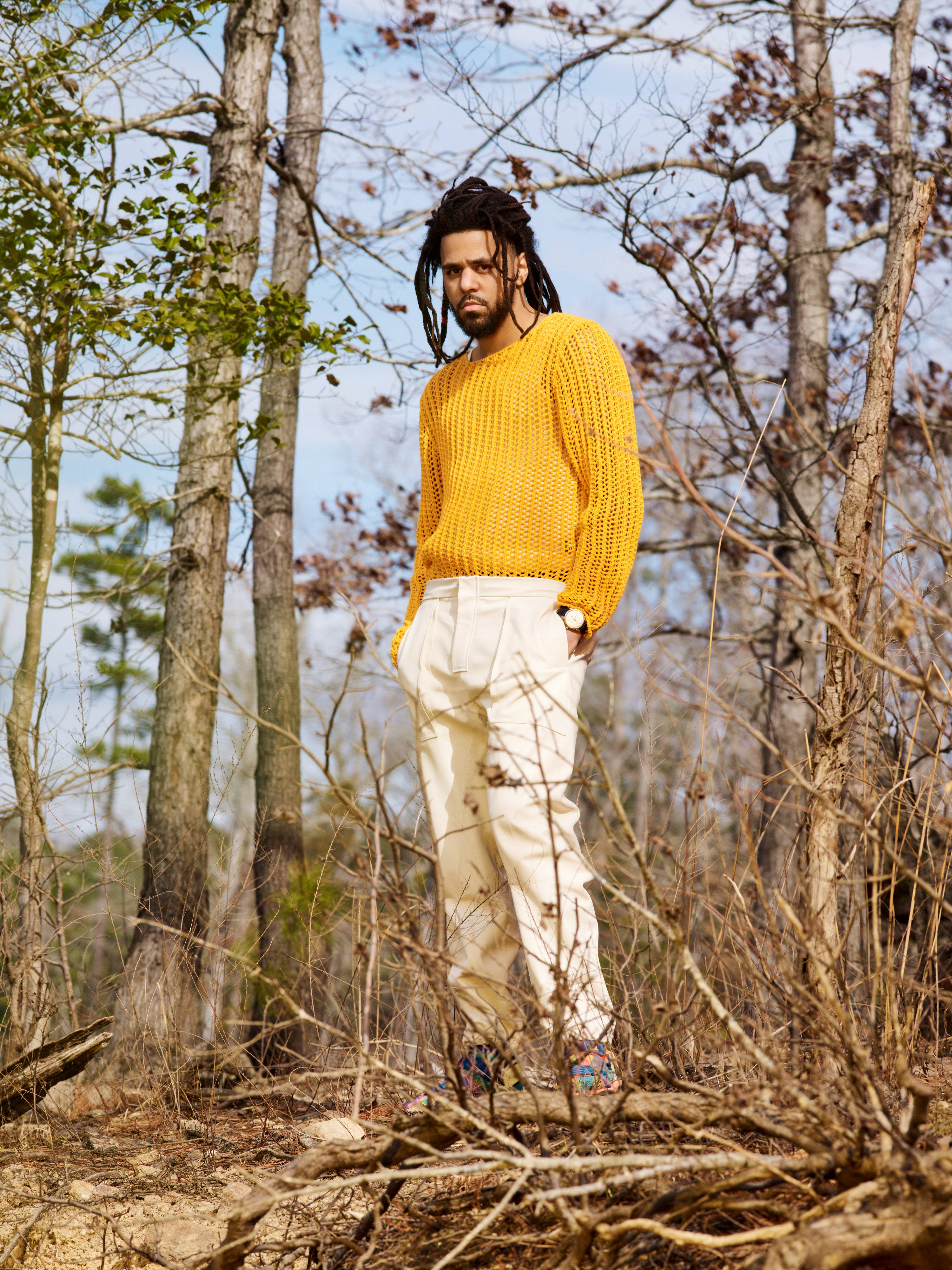 J Cole 黃色毛衣造型 壁紙 — 一名留著辮子、身穿亮黃色毛衣的男子自信地站在森林空地上的光禿樹木之間。