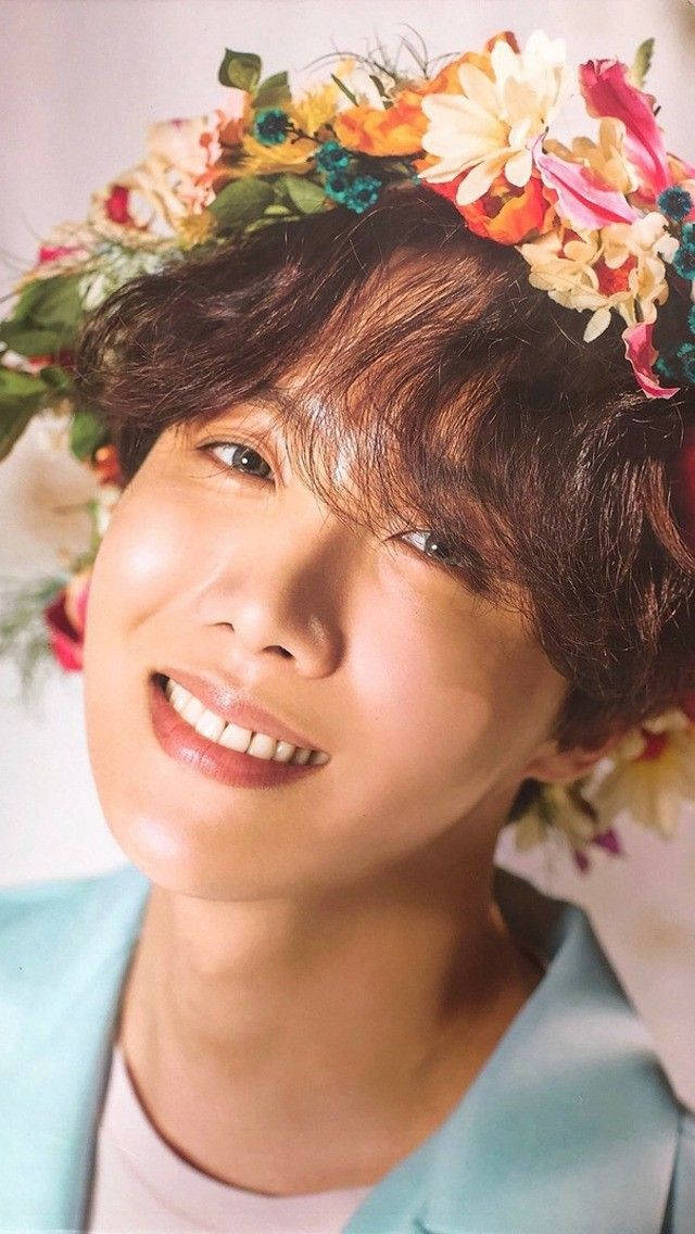 J-hope Flower Crown 壁紙 — 一位年輕男子帶著燦爛笑容，戴著色彩繽紛的花冠，背景是柔和的粉彩色。
